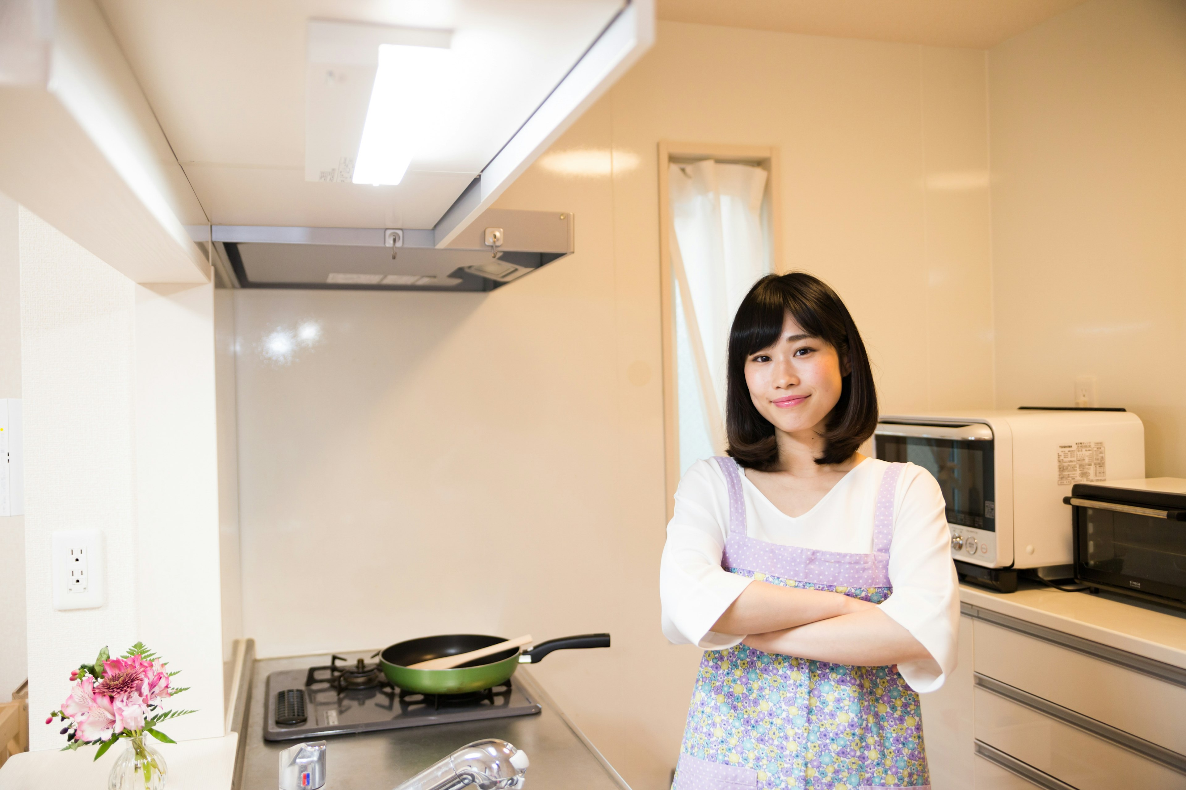 Wanita dengan tangan disilangkan di dapur cerah mengenakan atasan putih dan apron ungu dengan peralatan memasak dan bunga terlihat