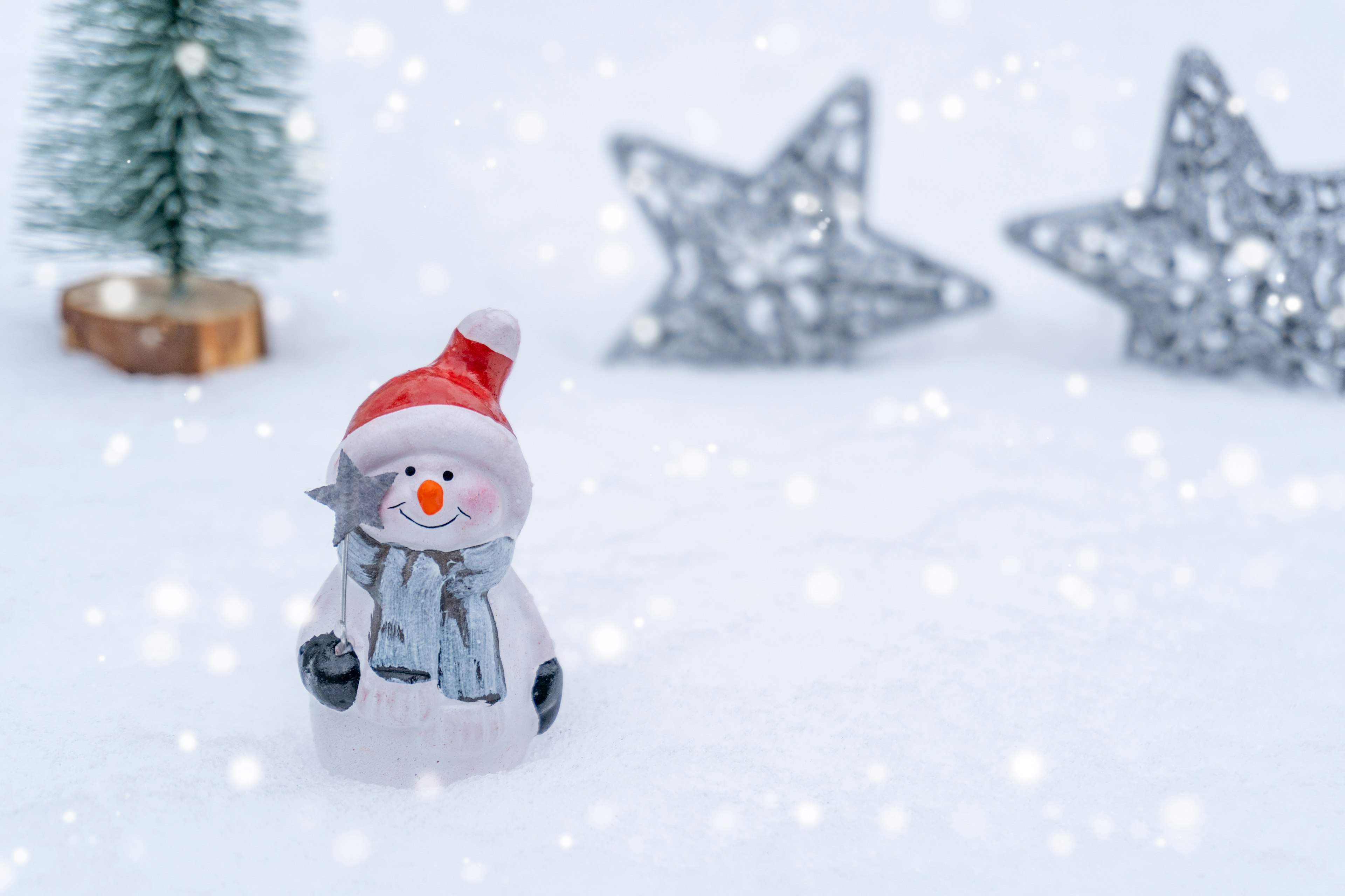 Snowman figurine on snow wearing a red hat and blue scarf small Christmas tree and silver stars in the background