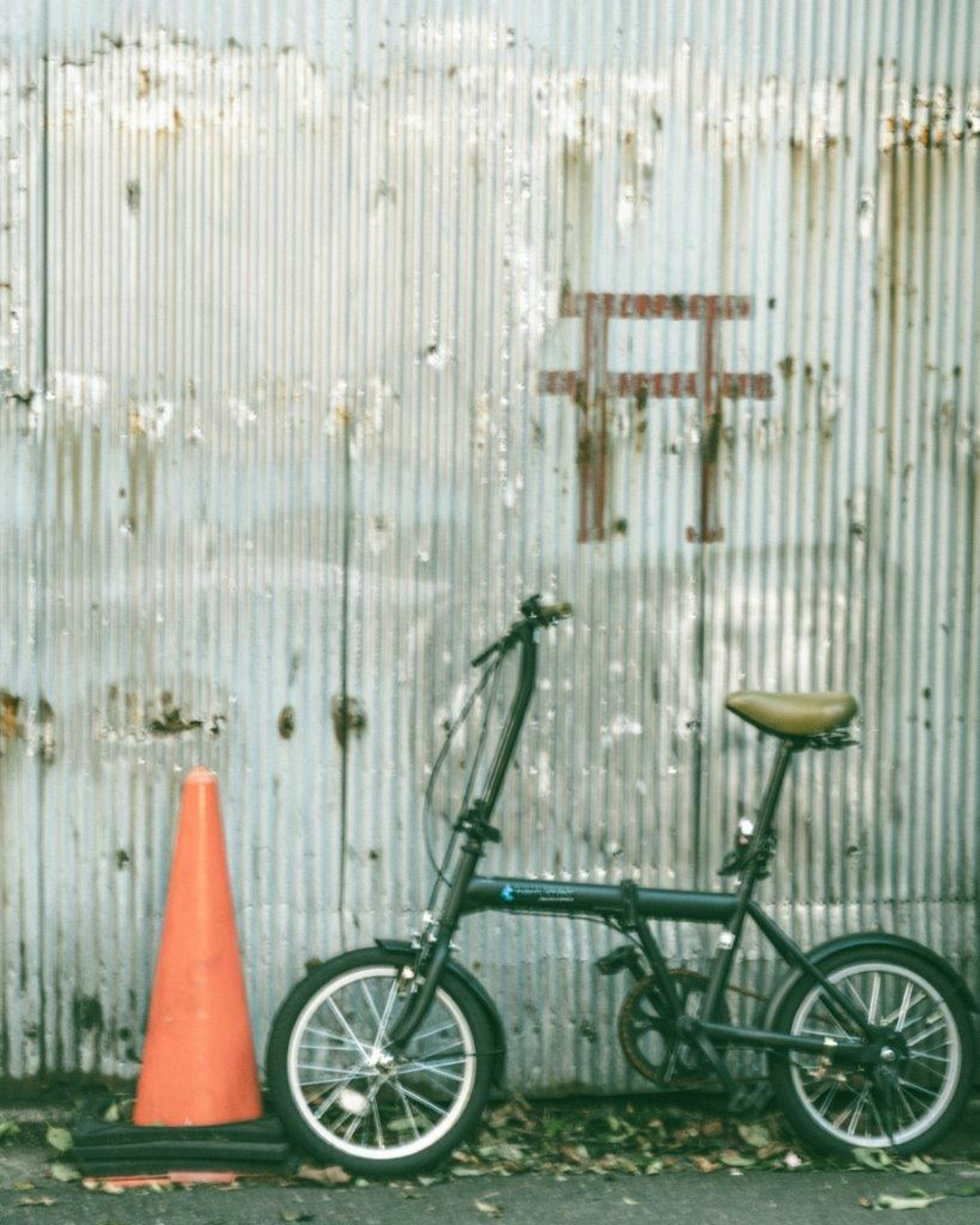 Foldable bicycle and orange traffic cone against a rusty metal wall