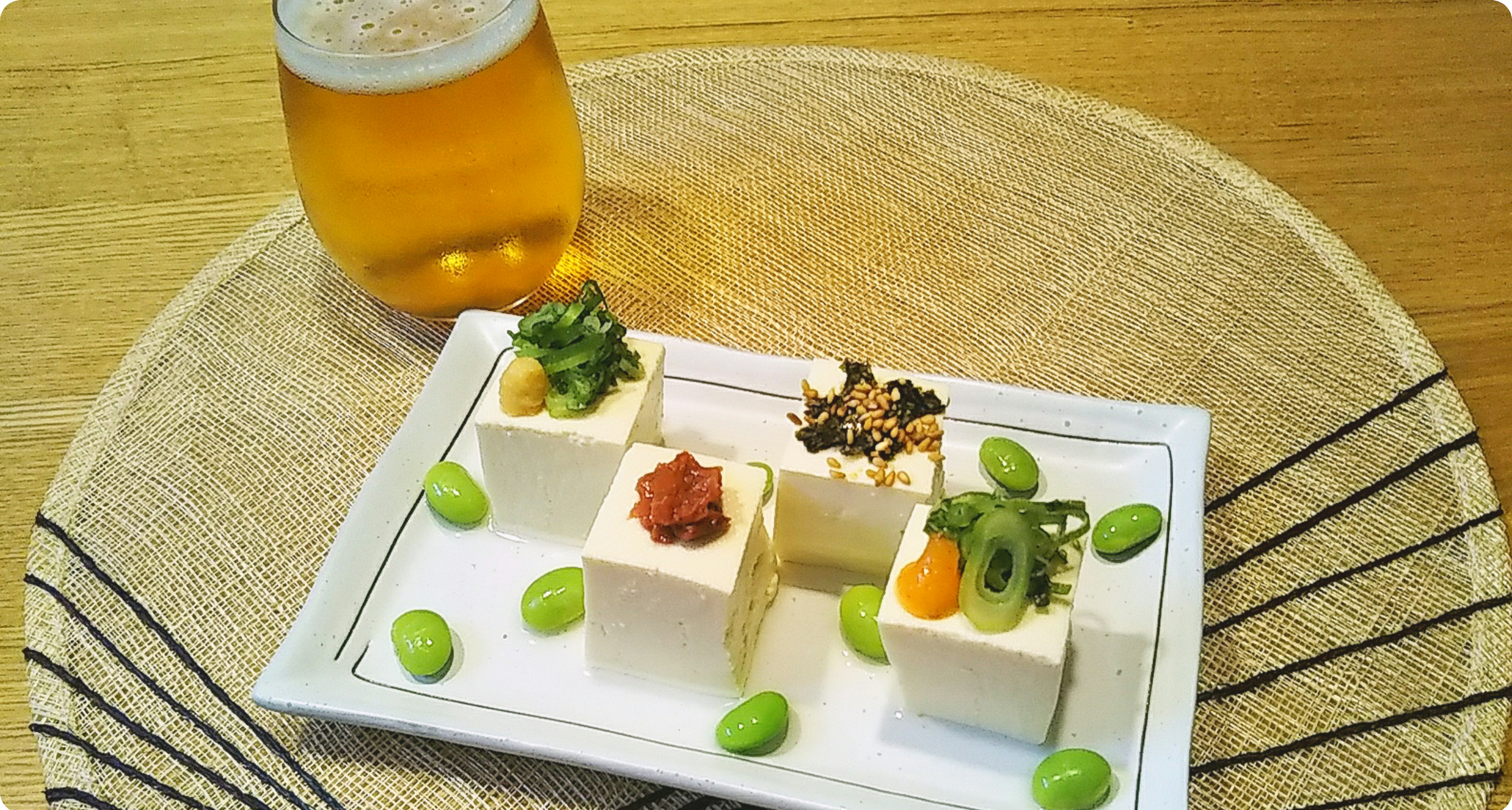 Plato de tofu decorado con coberturas coloridas y edamame junto a un vaso de cerveza