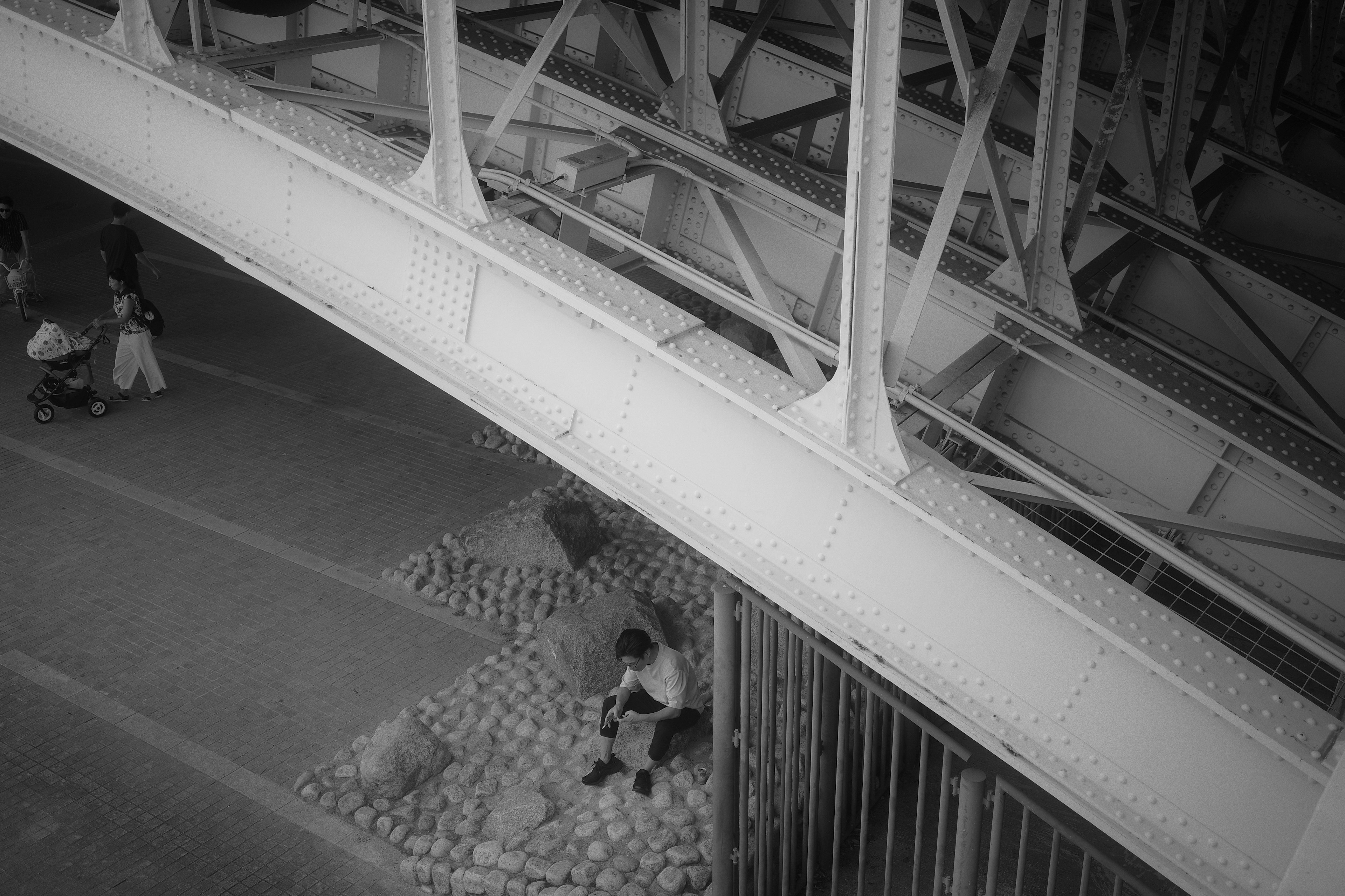 Zwei Personen sitzen unter einer schwarz-weißen Brücke mit Passanten