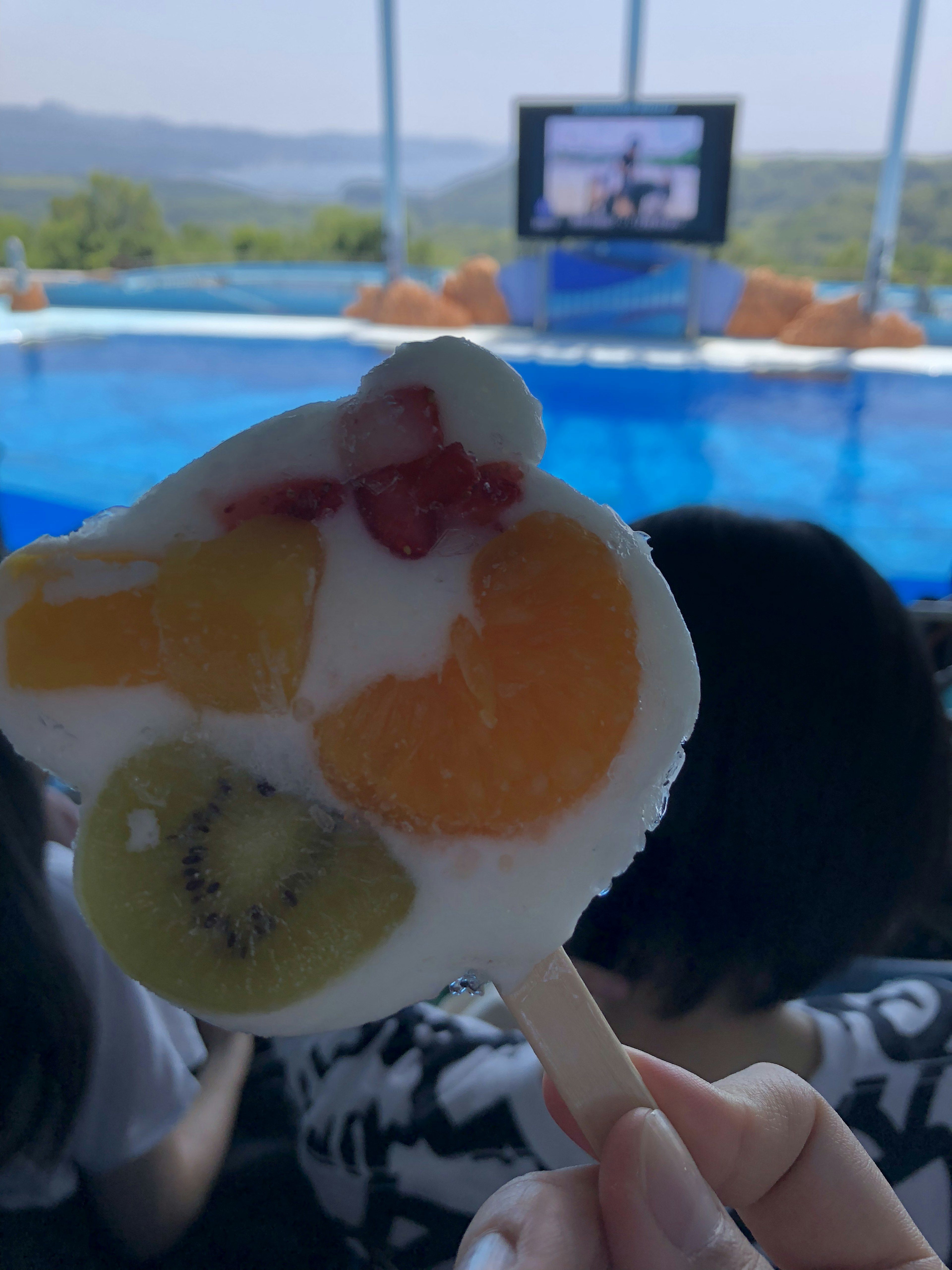 Hand holding a fruit-shaped ice pop in front of a pool