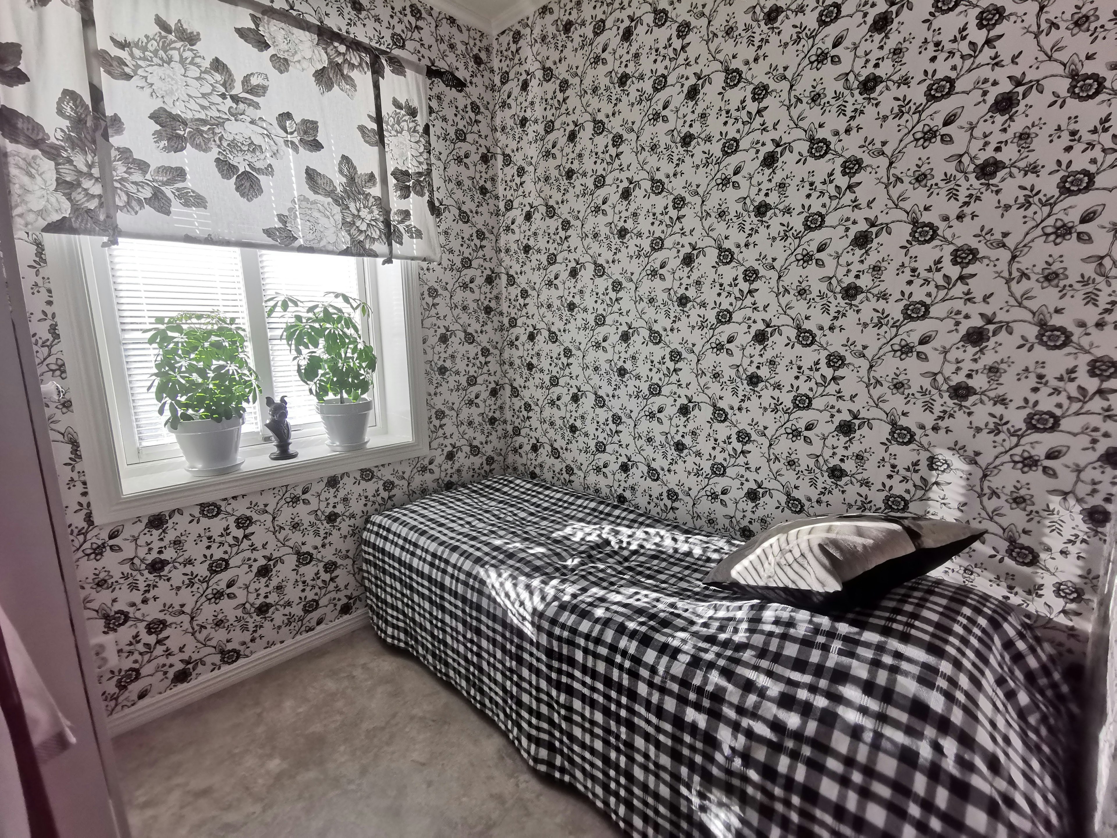 Pequeña habitación con papel tapiz floral en blanco y negro Plantas verdes en el alféizar de la ventana y colcha a cuadros