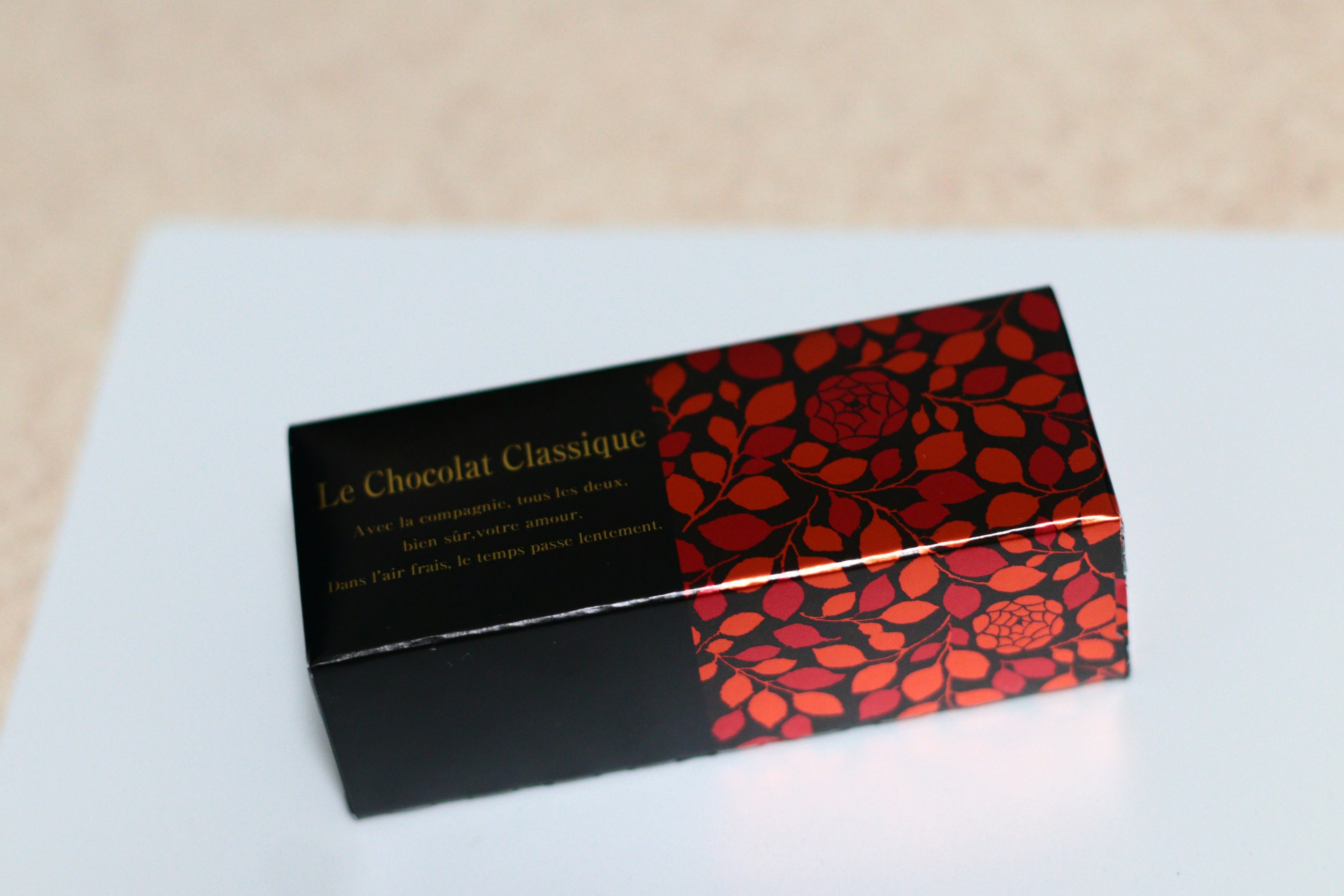 Caja negra con diseño floral rojo que presenta el texto 'Le Chocolat Classique'