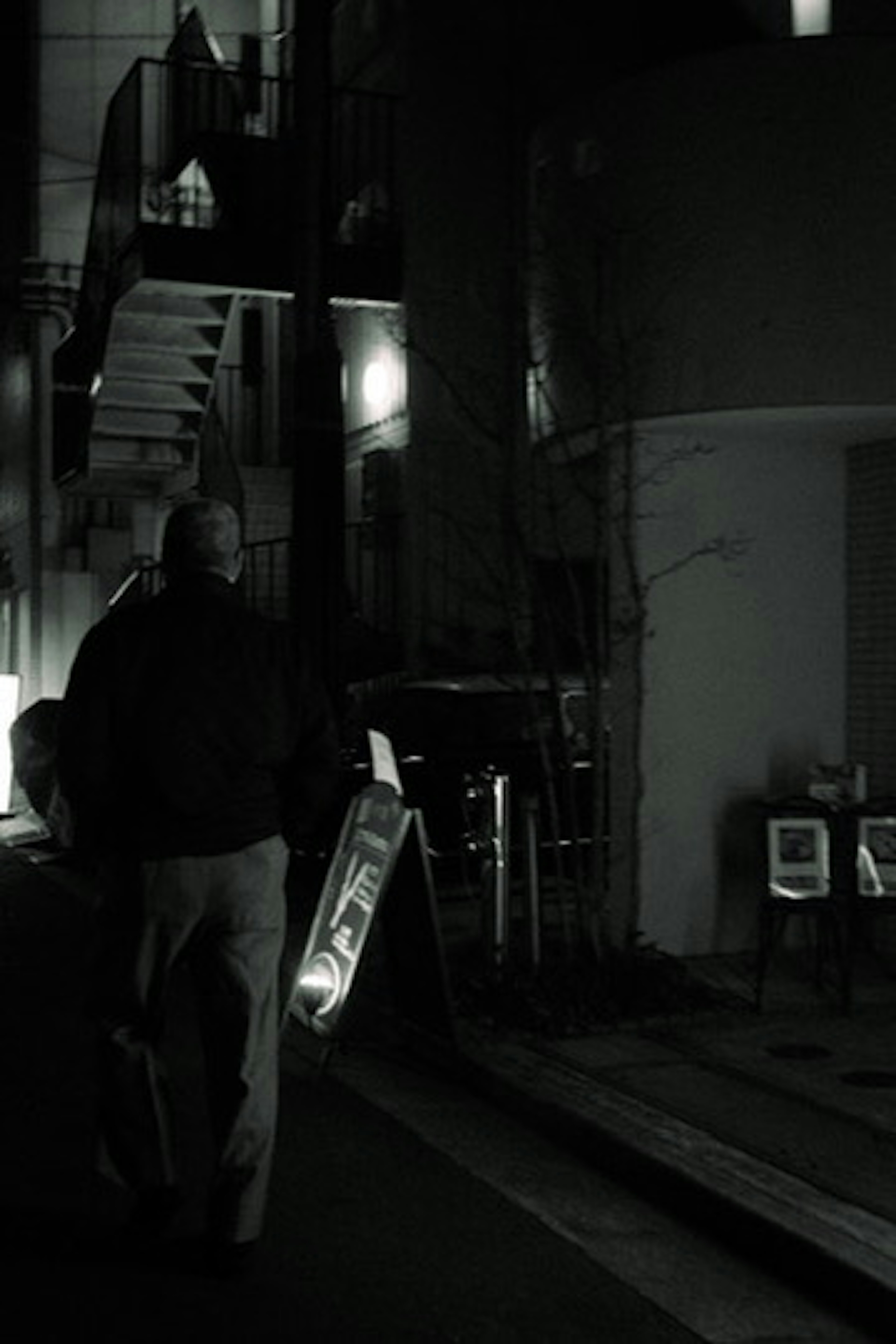 A figure walking in a dark alley with a building featuring stairs