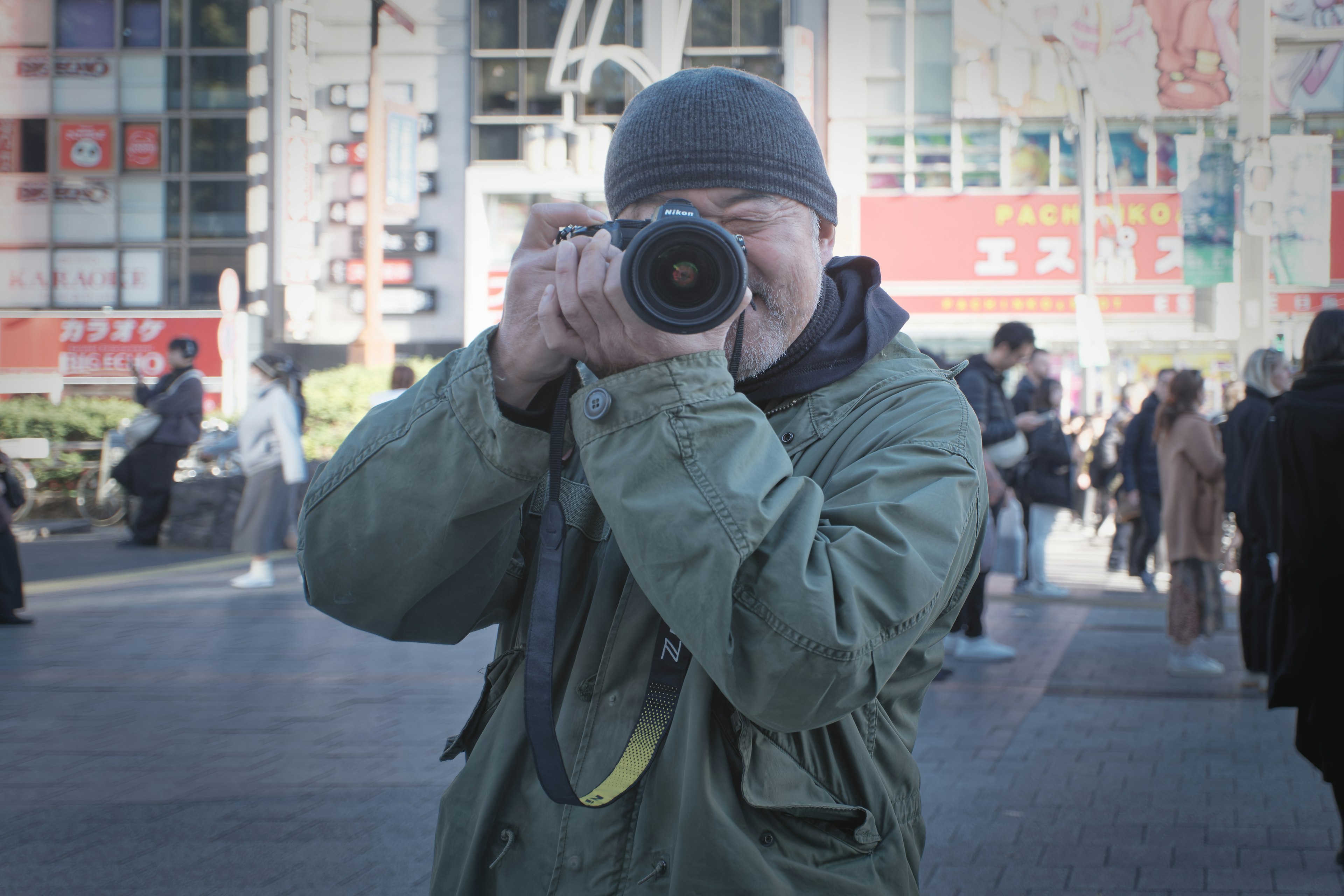 Uomo con una macchina fotografica che punta l'obiettivo verso l'osservatore persone che camminano sullo sfondo