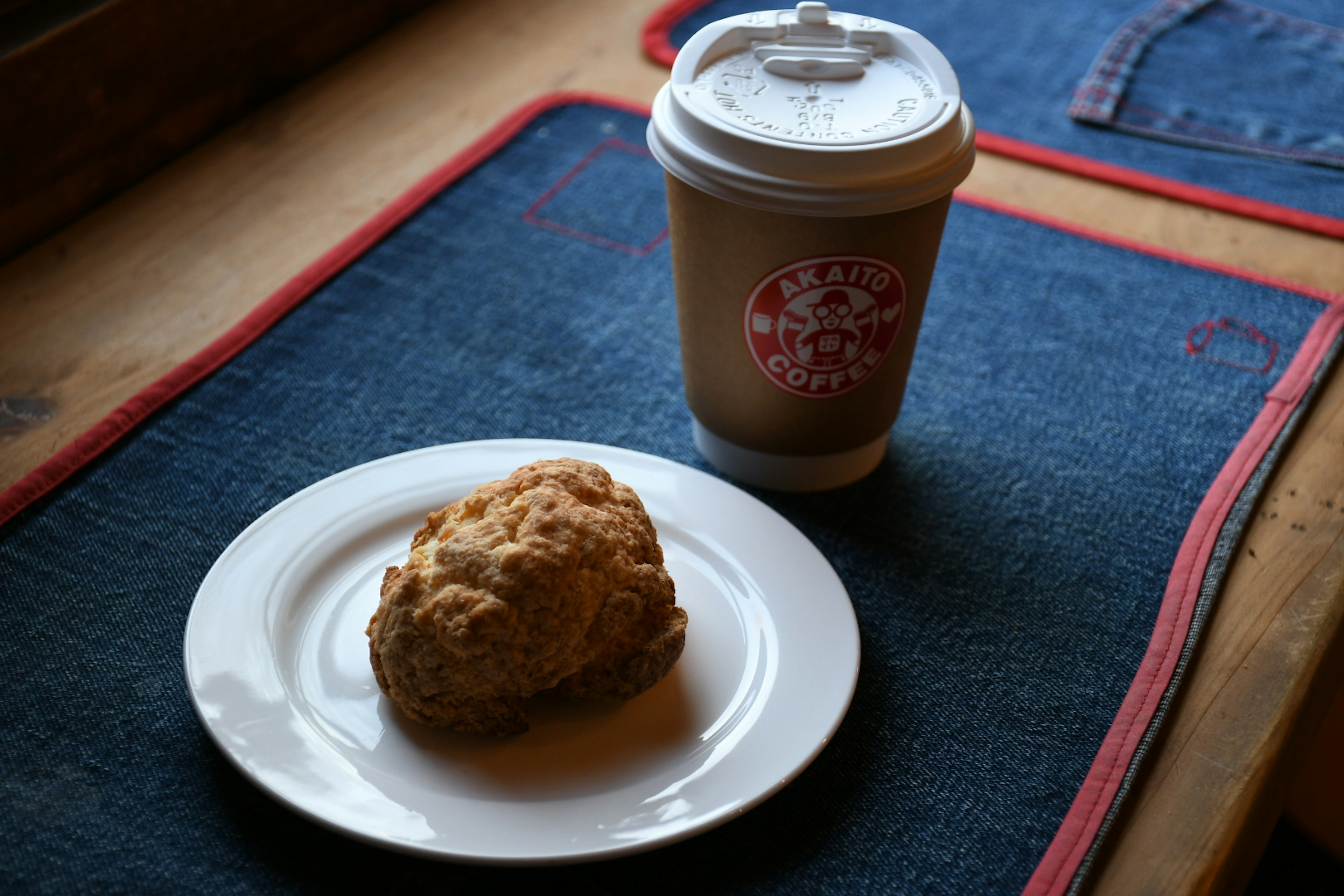 A biscuit on a white plate next to a coffee cup on a denim placemat