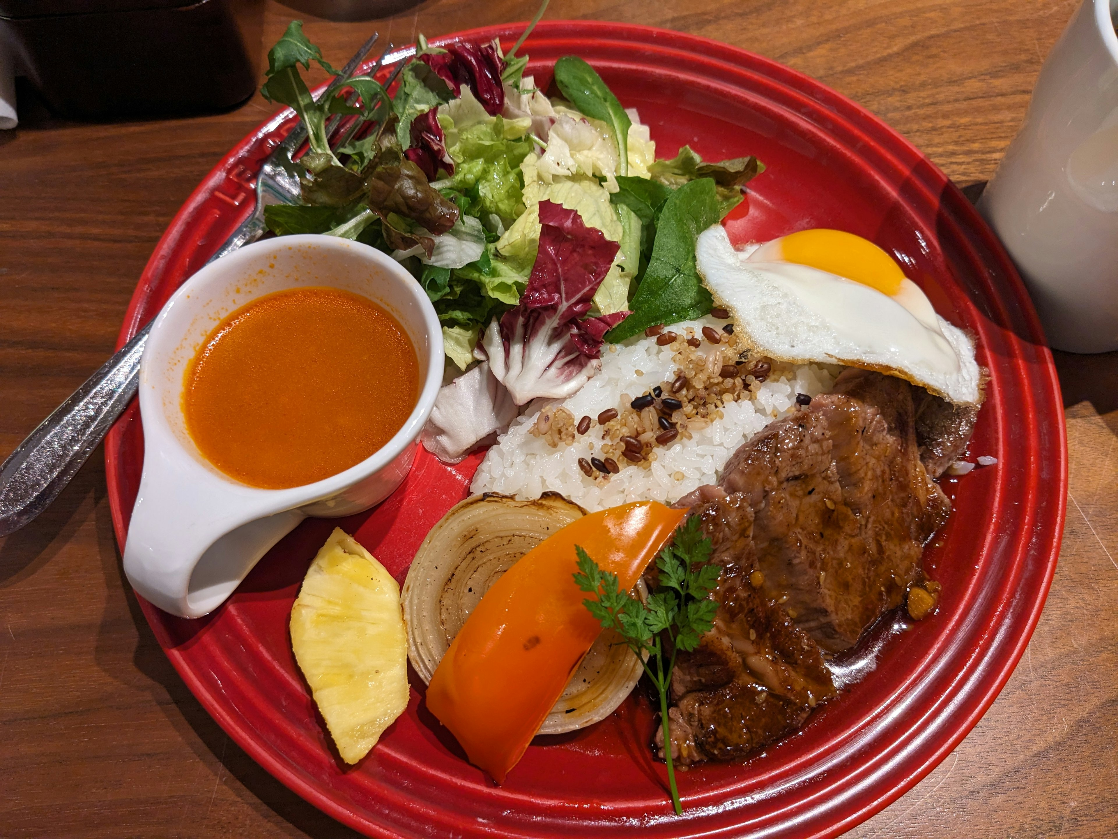 A vibrant red plate featuring a salad, a fried egg, meat, rice, and sauce