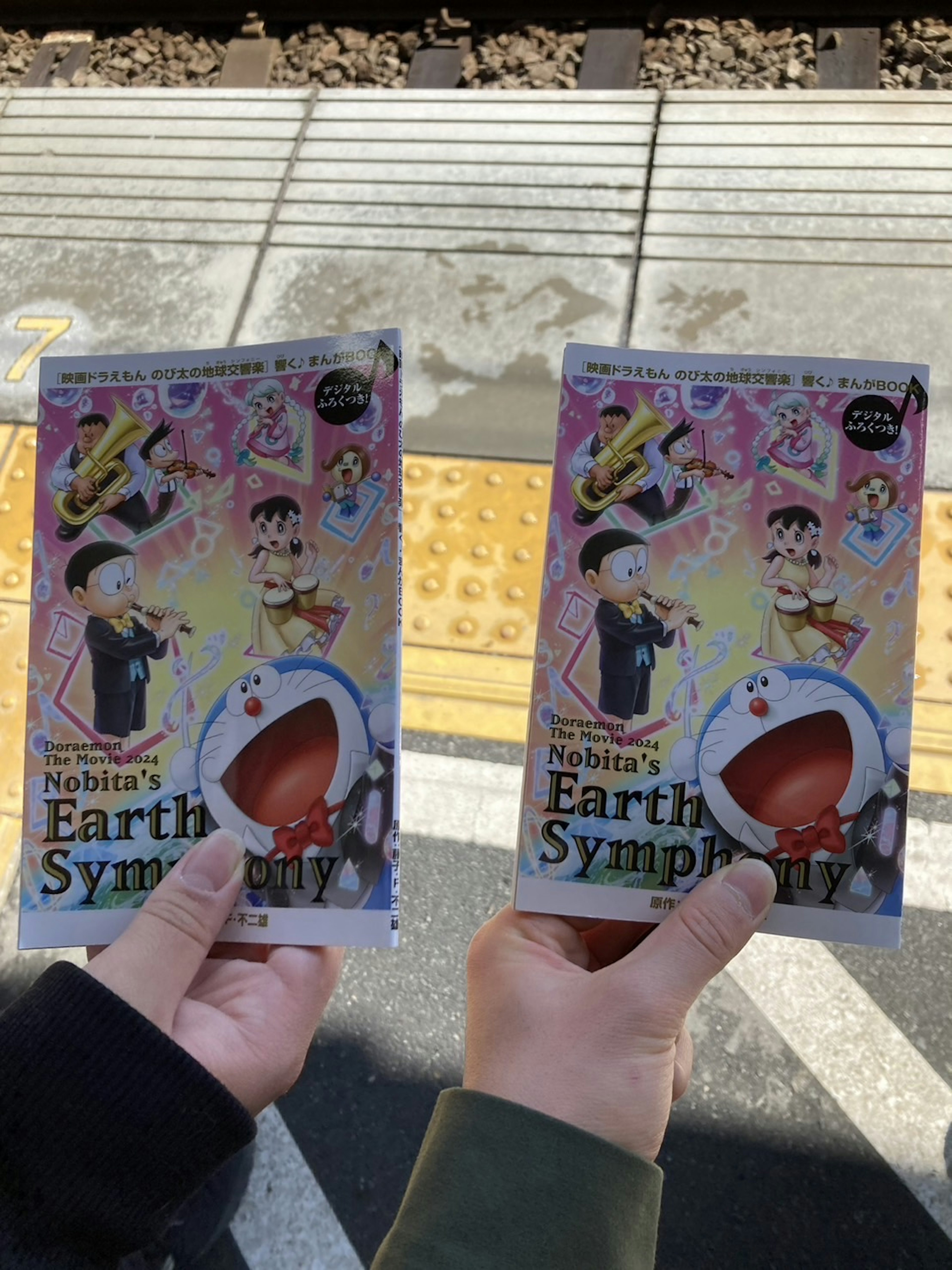 二人が持っている「Earth Symphony」の冊子が駅のプラットフォームで見える