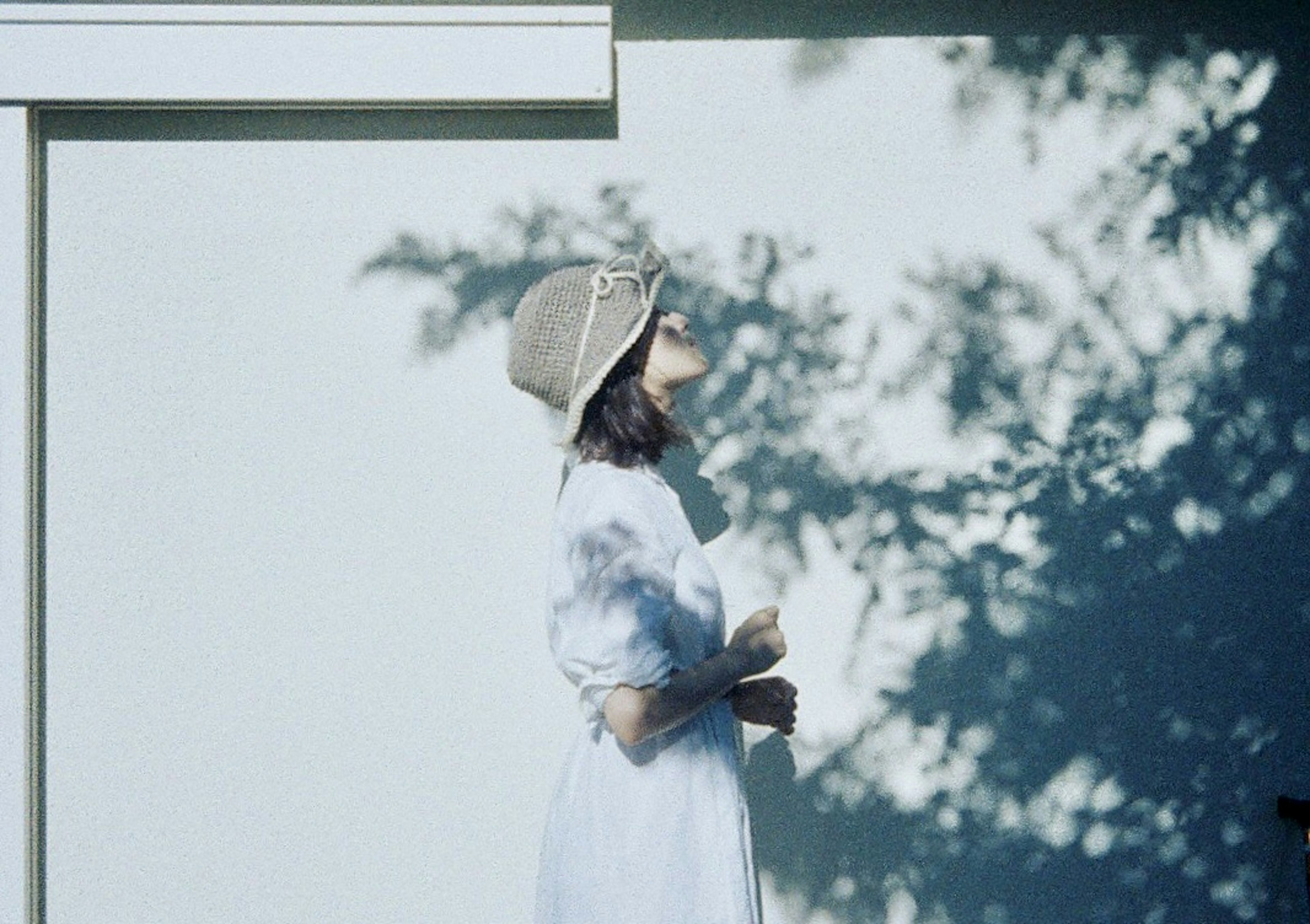 Une femme en robe blanche portant un chapeau se tient devant un mur avec des ombres