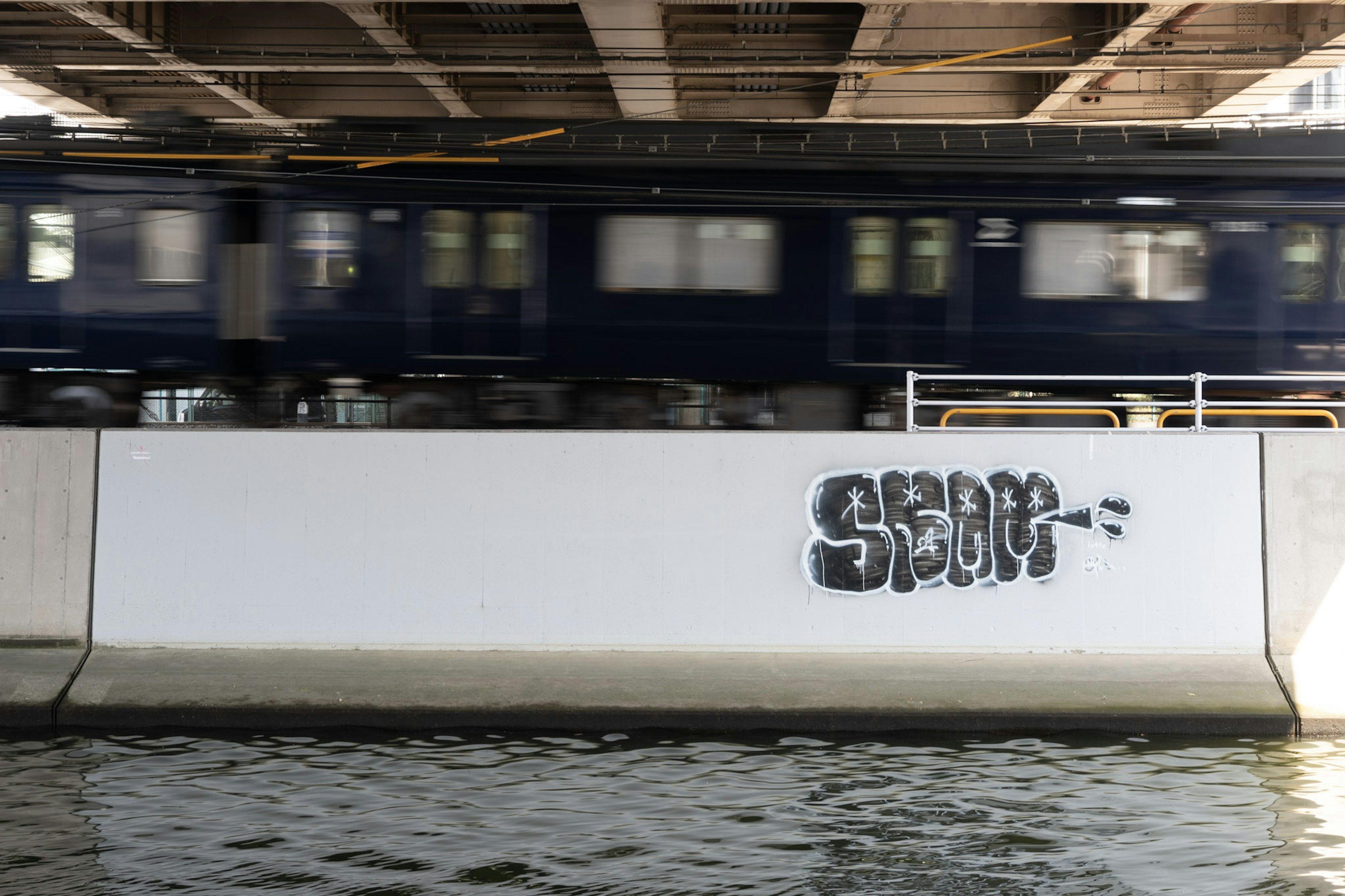 Train bleu passant sous un pont avec graffiti sur un mur blanc