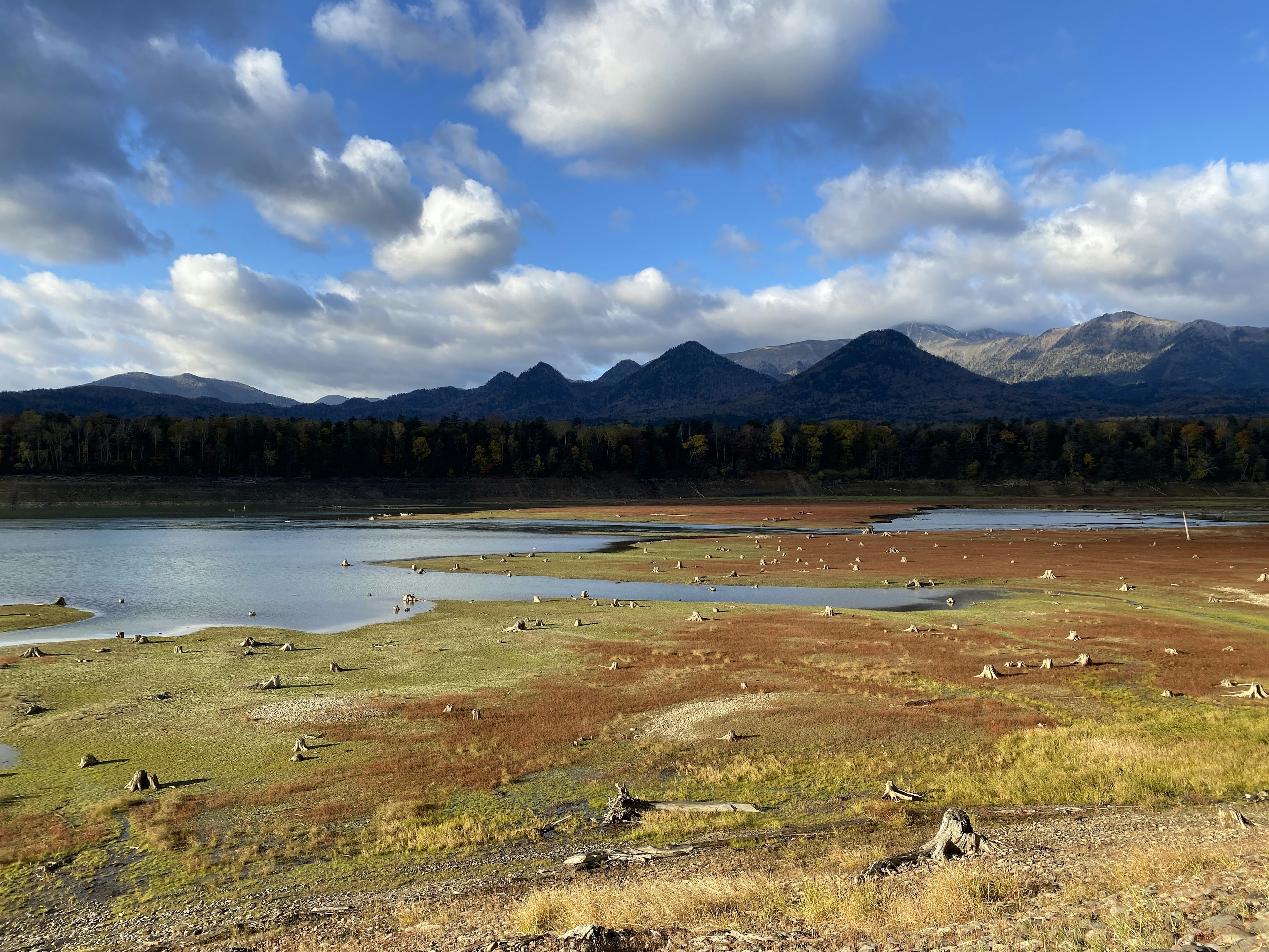 青空と雲に囲まれた湖の風景、遠くの山々と乾燥した土、岩が点在する