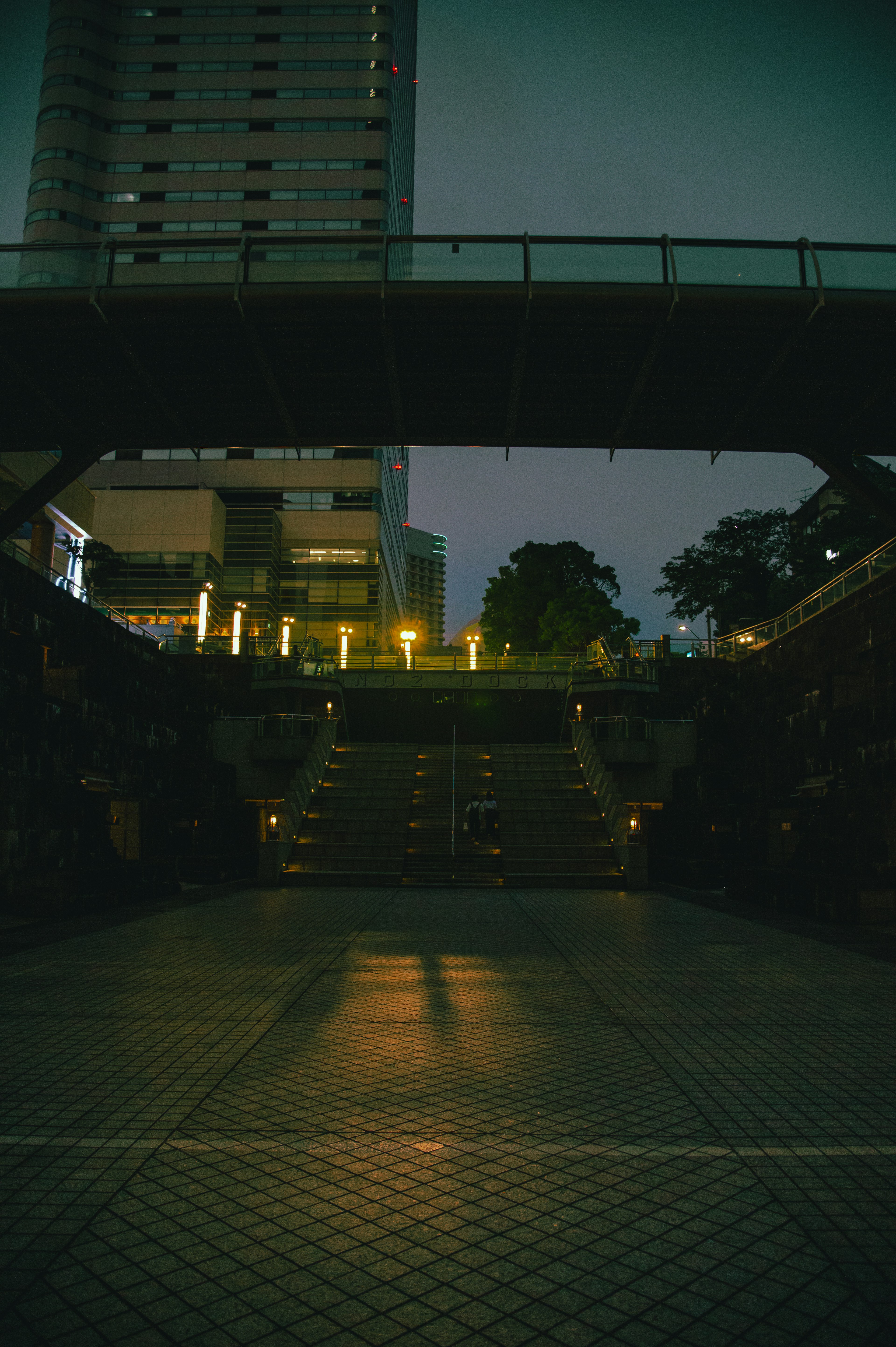 Scène urbaine nocturne sous un pont avec des escaliers illuminés et de la verdure