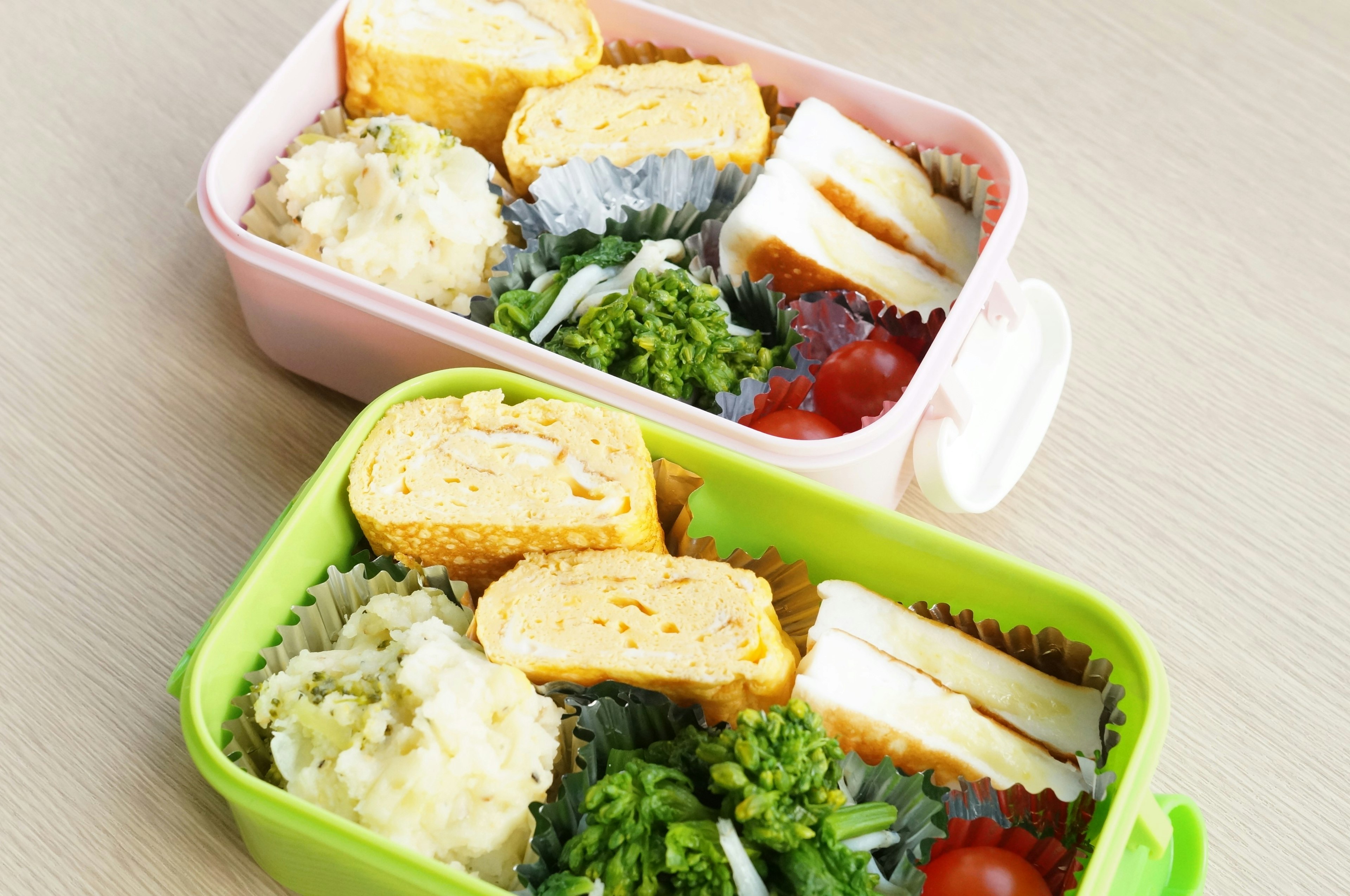 Pink and green bento boxes filled with tamagoyaki, broccoli, potato salad, and cherry tomatoes