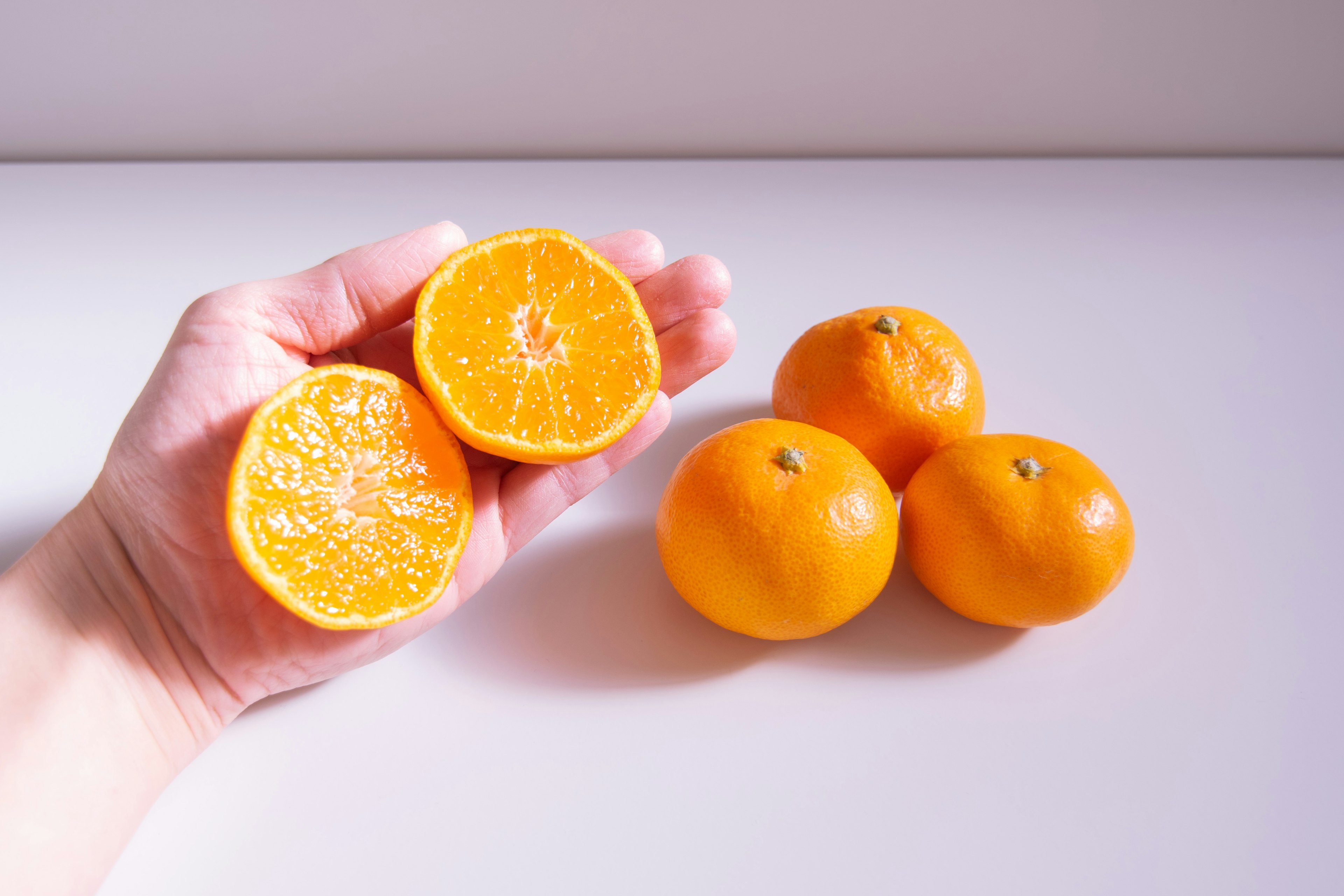 Eine Hand hält eine halbierte Orange, während vier ganze Orangen auf einer weißen Oberfläche liegen