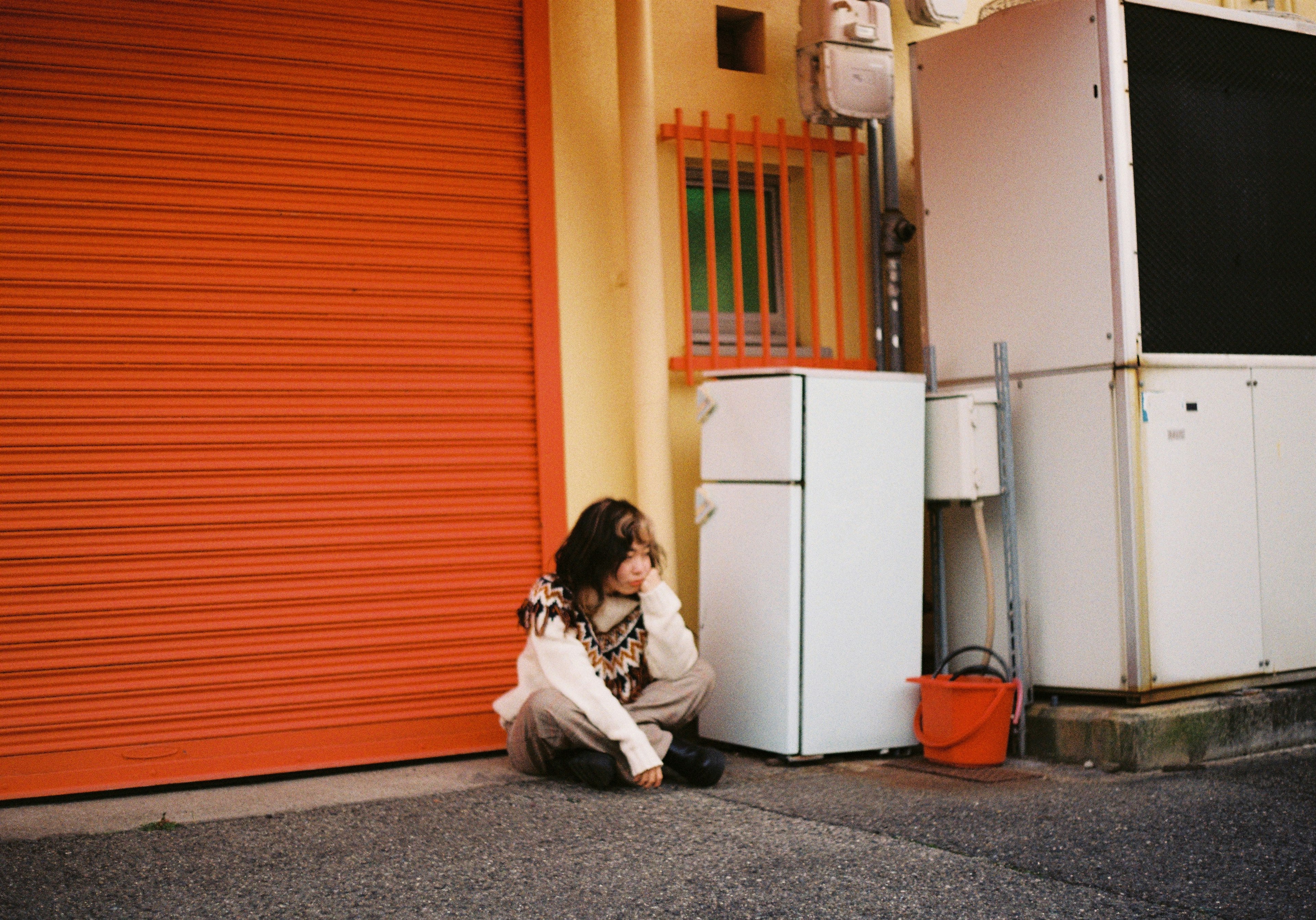 Femme assise devant un rideau orange avec un réfrigérateur blanc et un seau orange à proximité