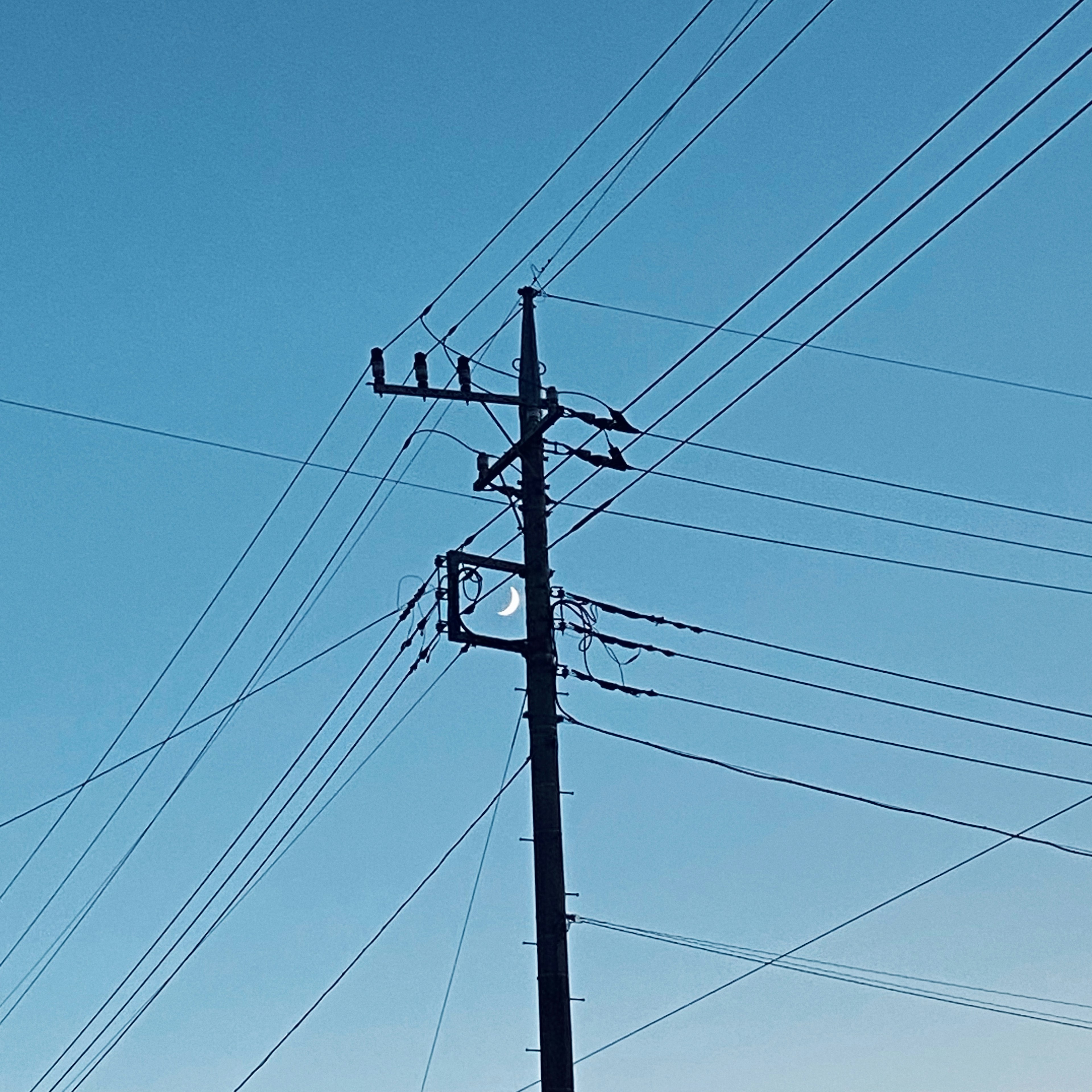 Silueta de la luna enmarcada por un poste de luz y cables bajo un cielo azul