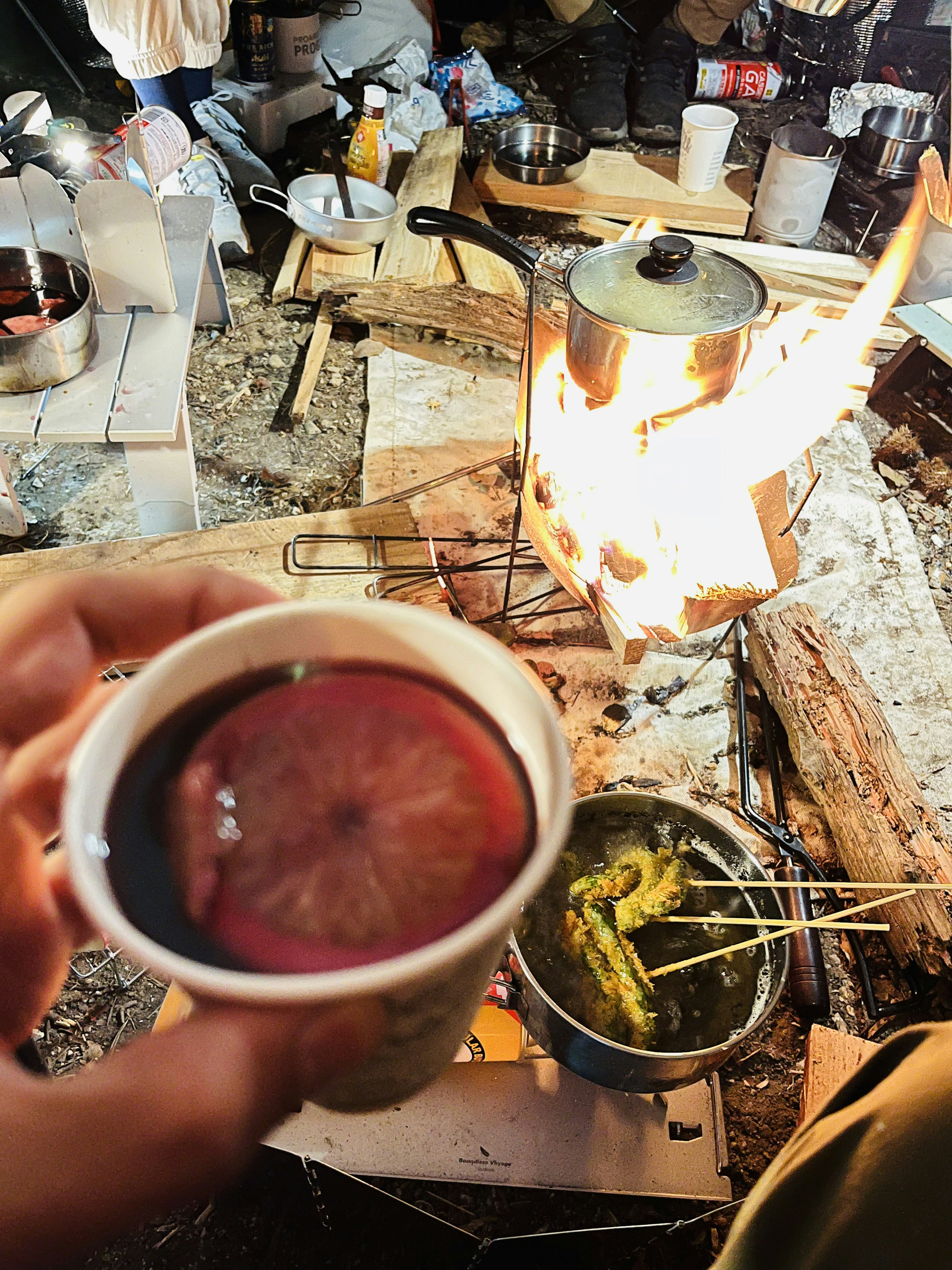 A hand holding a cup of warm drink with a pot cooking over a fire in the background