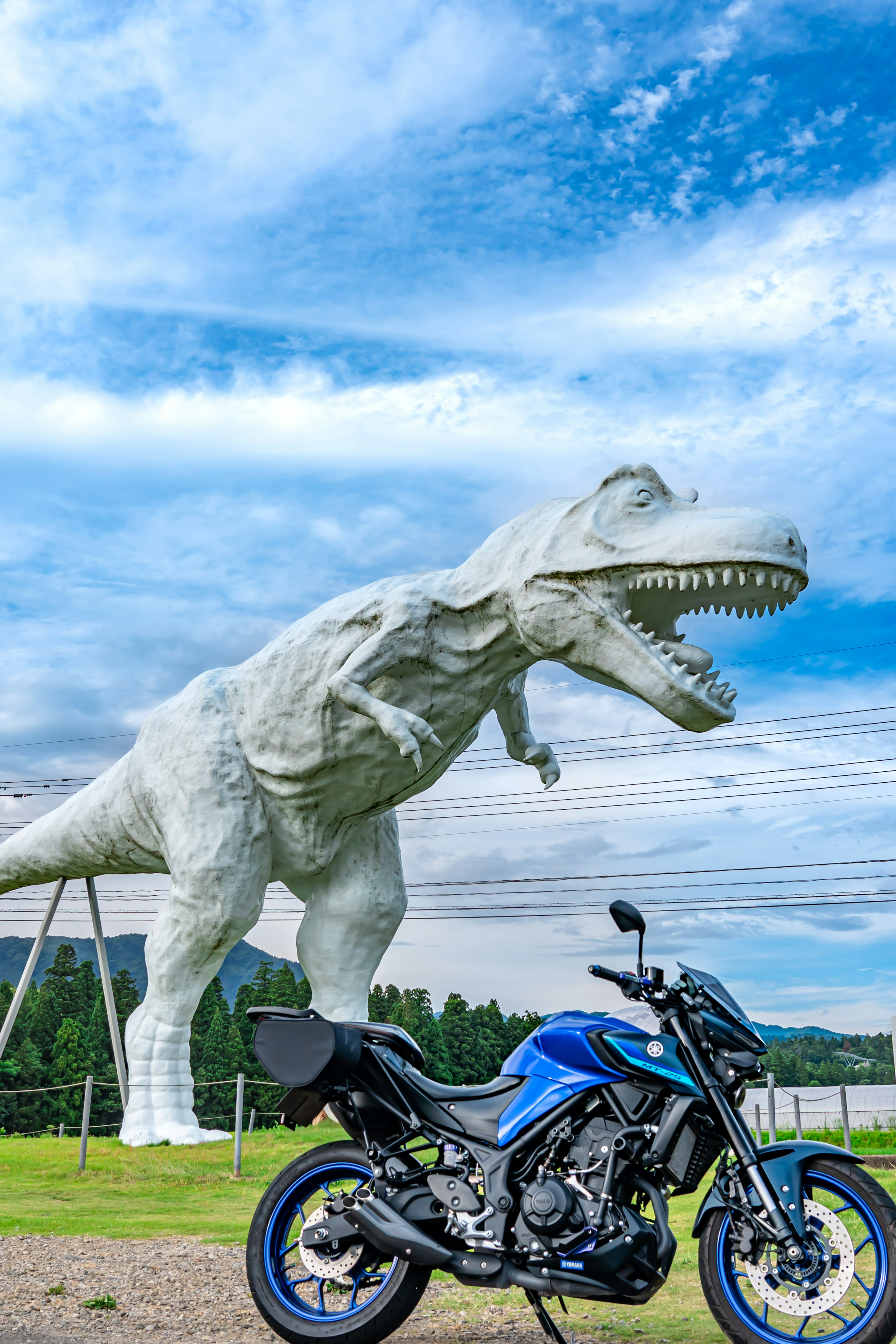 Una escultura gigante de dinosaurio blanco junto a una motocicleta azul bajo un cielo azul