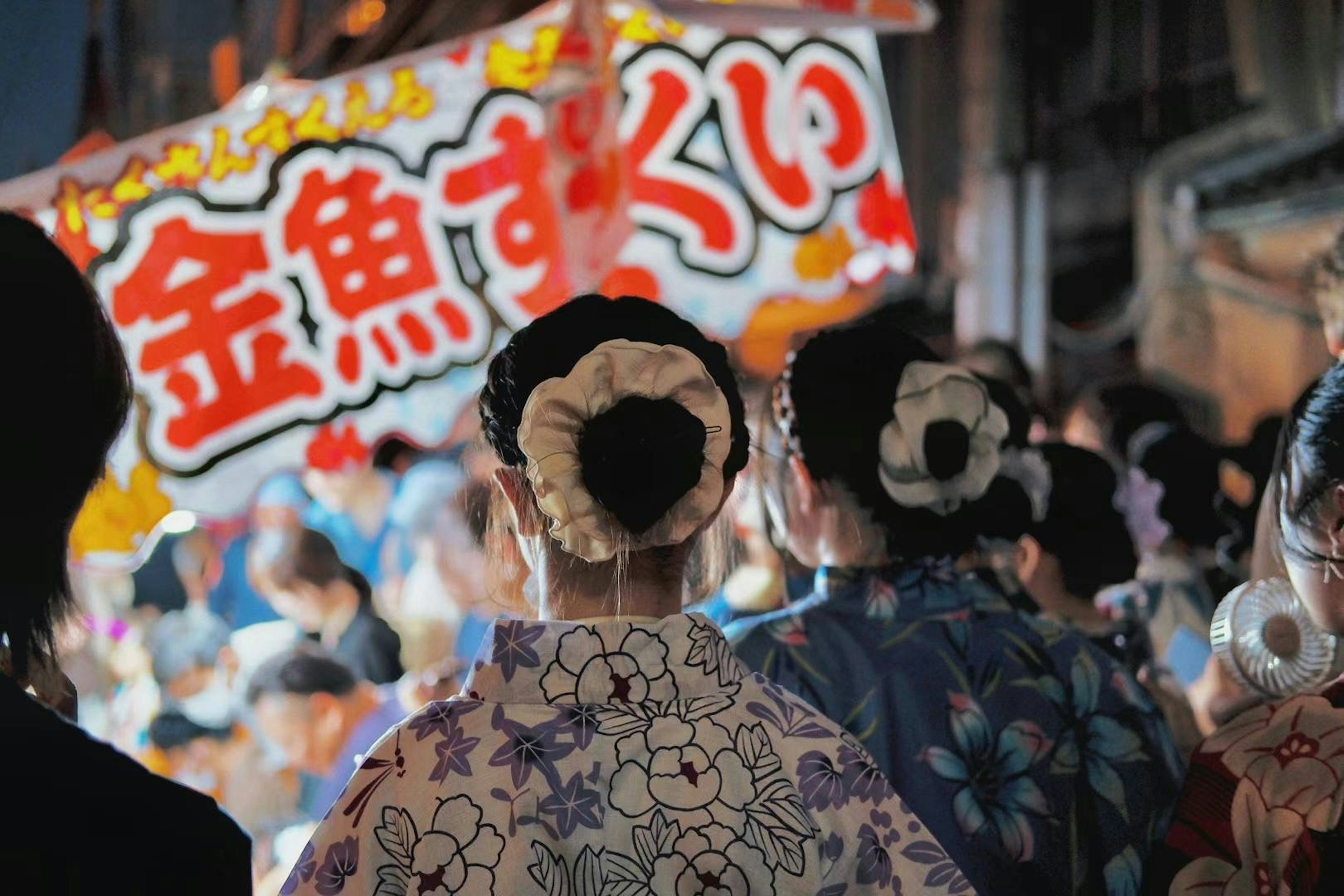 夜の祭りで着物を着た女性たちの後ろ姿と金魚すくいの看板