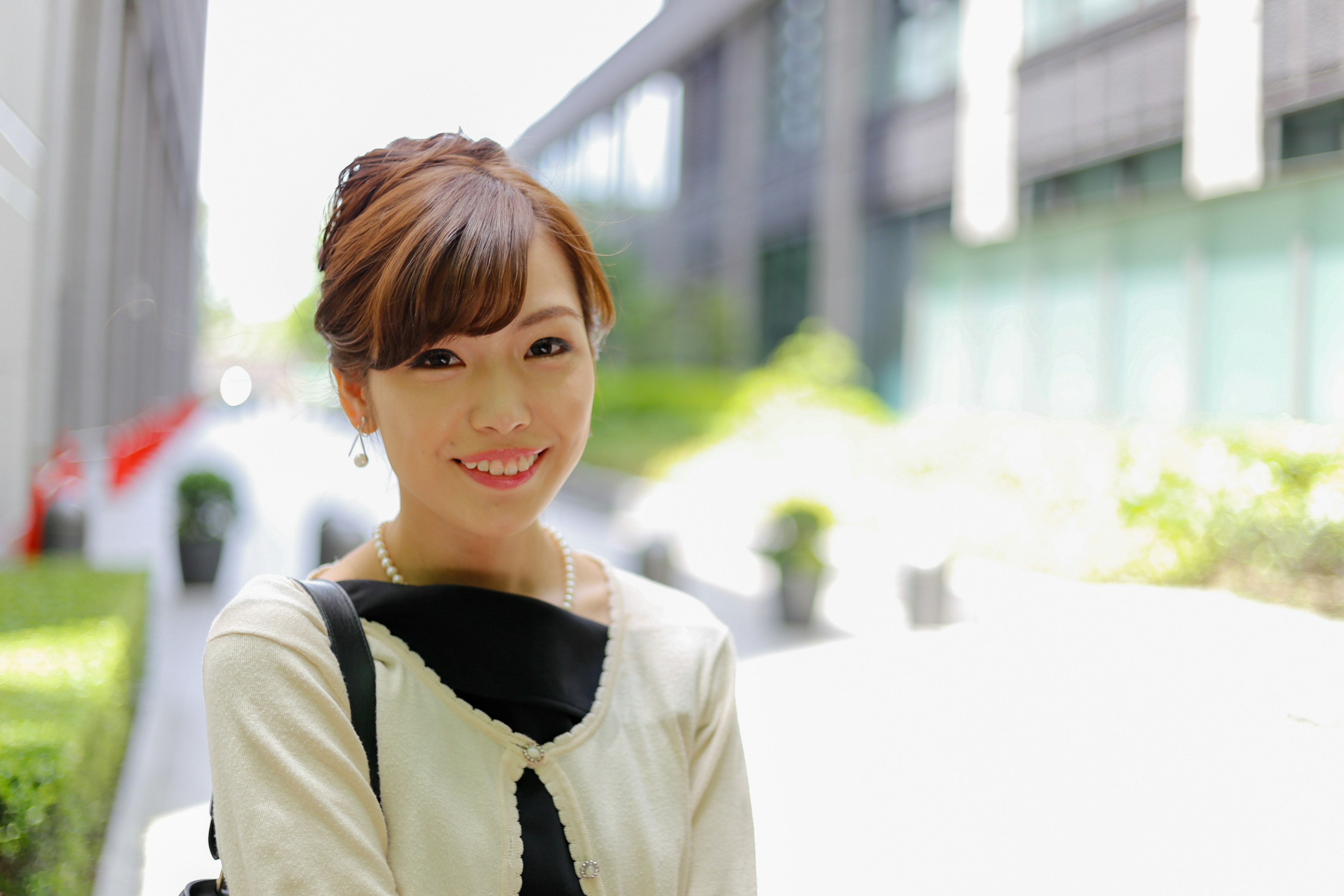 A woman smiling in an outdoor scene with buildings and greenery in the background