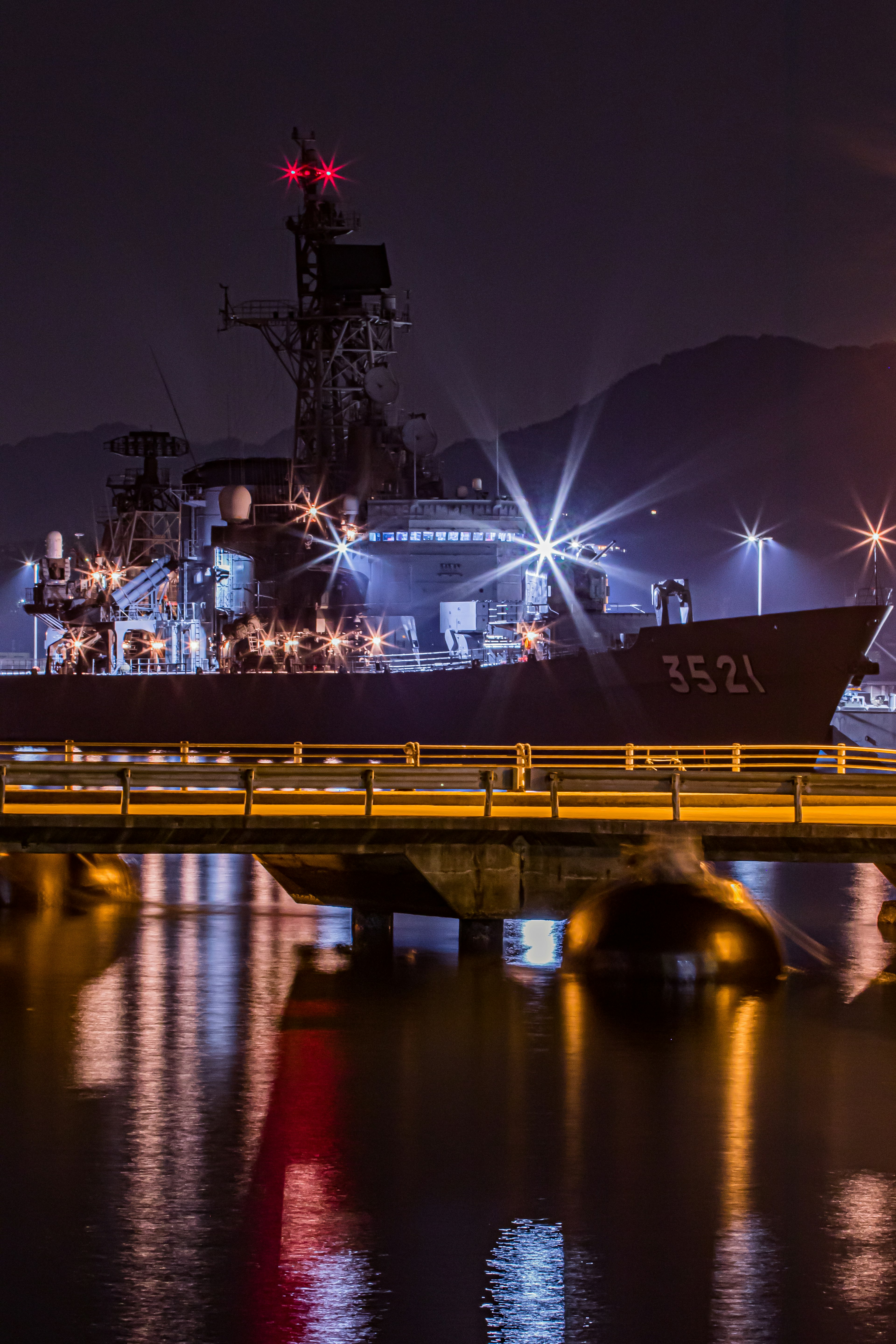 A naval ship docked at night with bright lights reflecting on the water