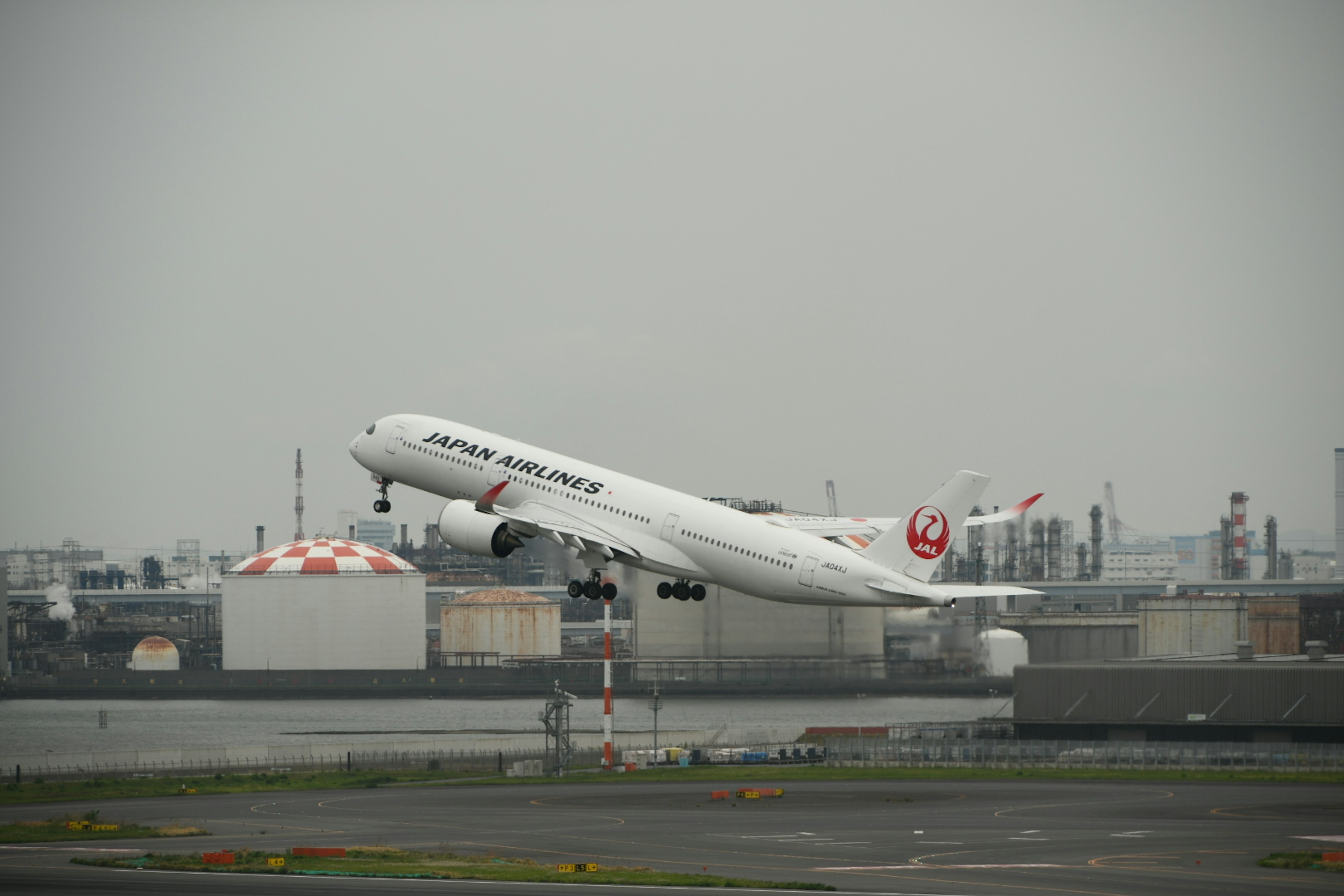 เครื่องบินโดยสาร Japan Airlines ขึ้นบินท่ามกลางท้องฟ้าสีเทาและฉากหลังอุตสาหกรรม