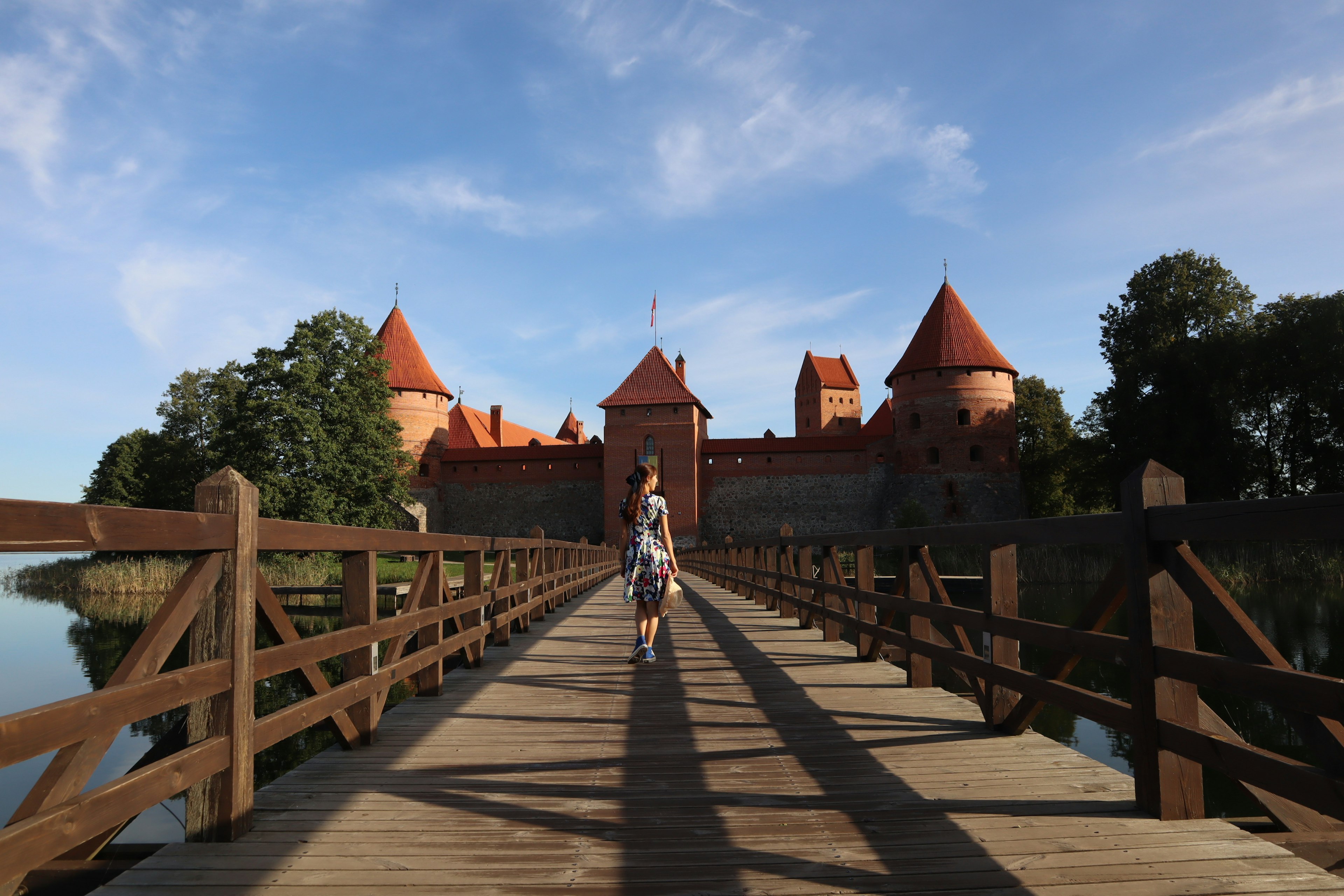 Una mujer caminando por un puente de madera sobre un lago con un castillo de techo rojo al fondo