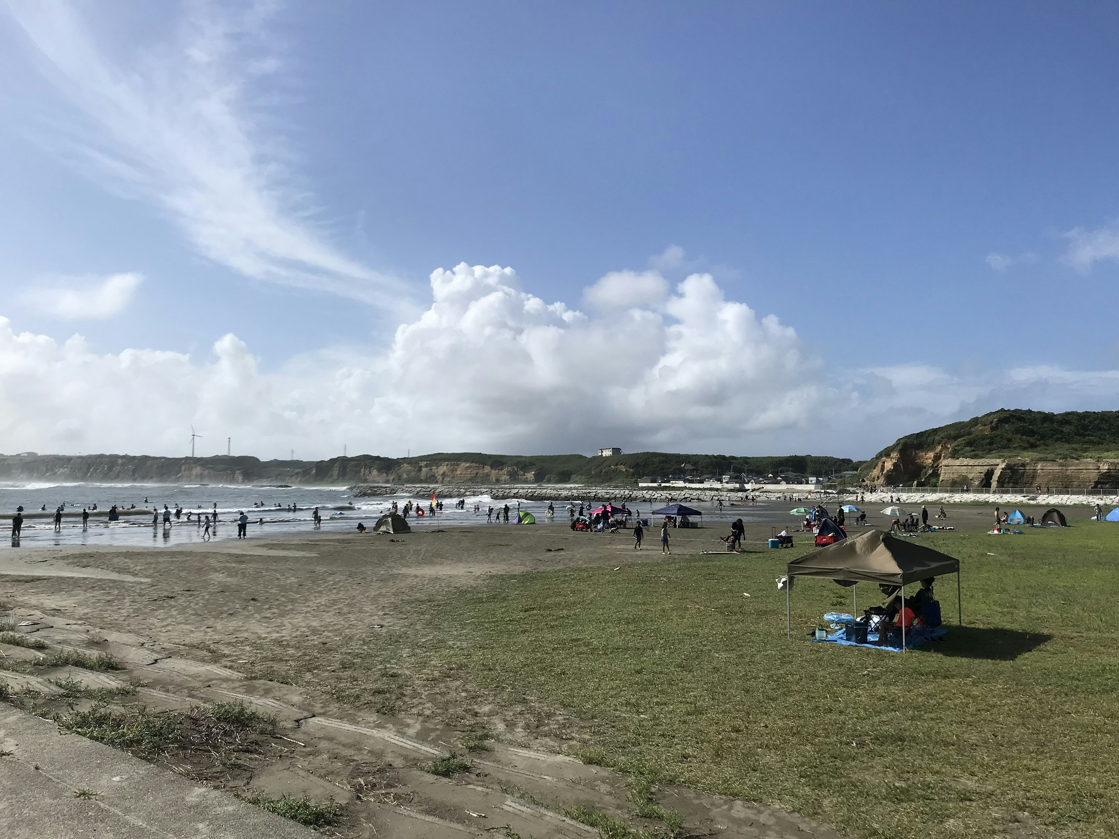 Escena de playa con personas disfrutando del agua bajo un cielo azul y nubes esponjosas