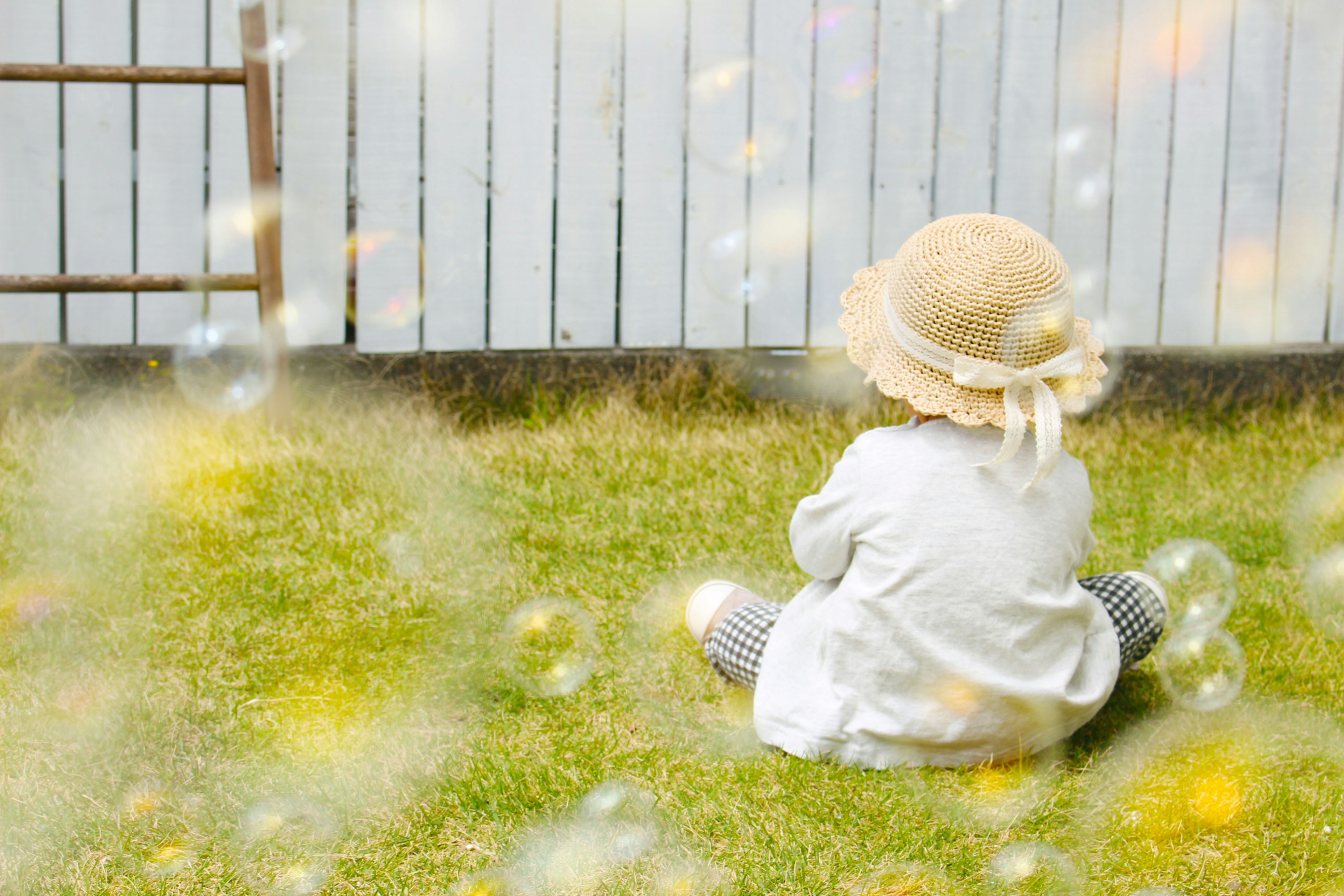 A child in a white outfit and straw hat sitting on grass with bubbles floating around