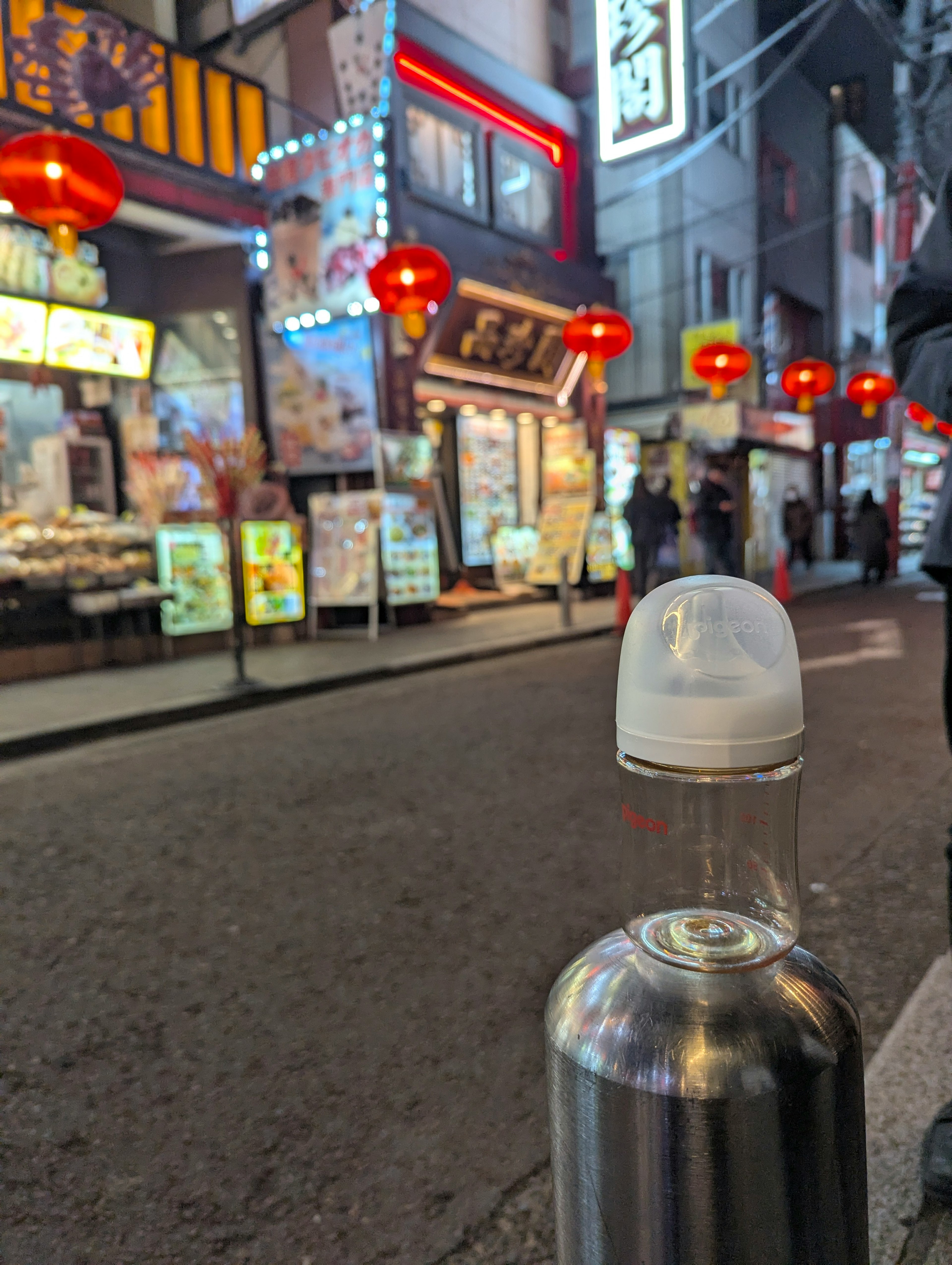 Scena notturna di strada con una bottiglia d'acqua con cappuccio bianco e lanterne rosse che illuminano i negozi