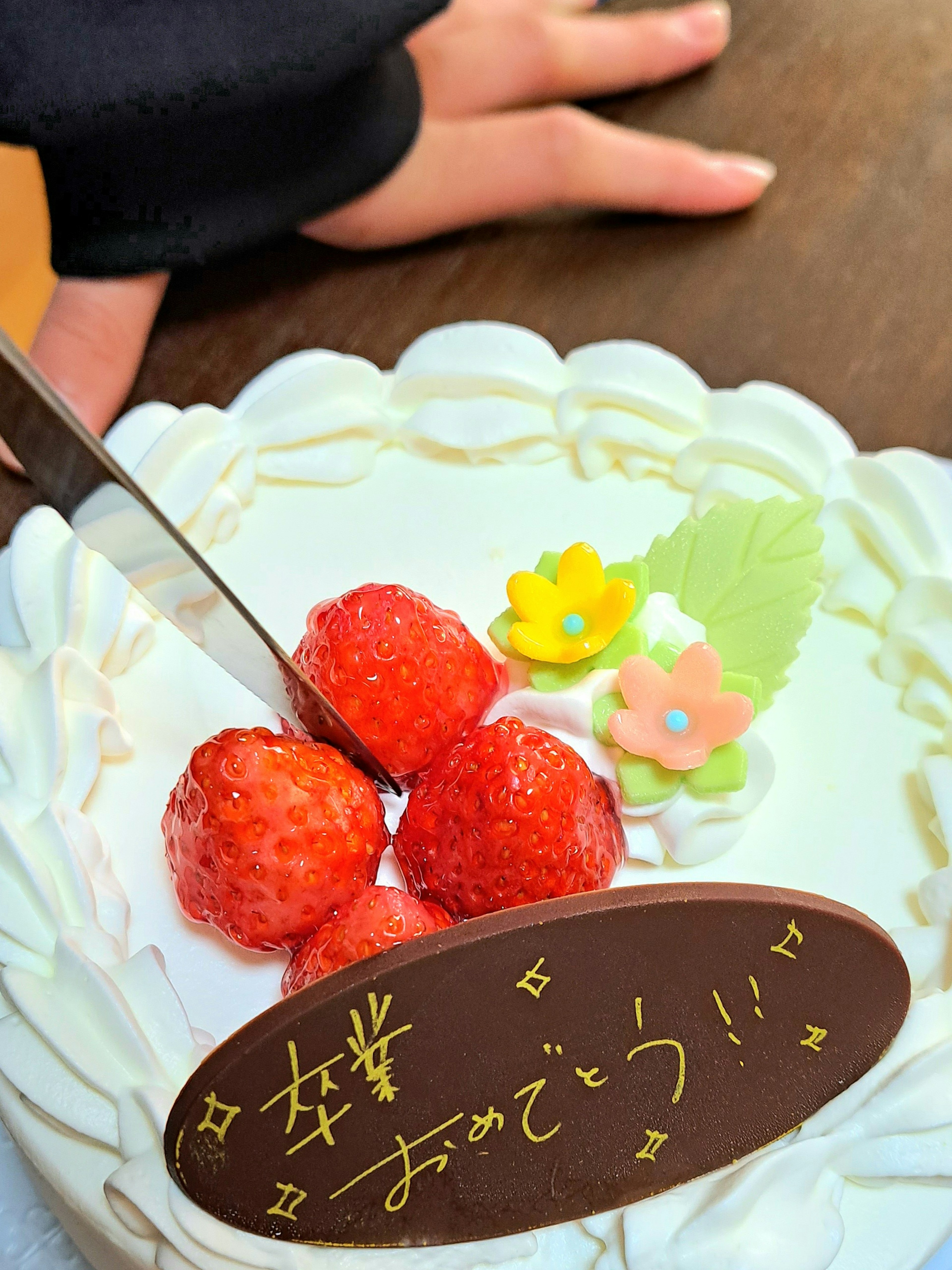 Un pastel de crema blanca decorado con fresas y flores con un plato de chocolate que dice felicidades