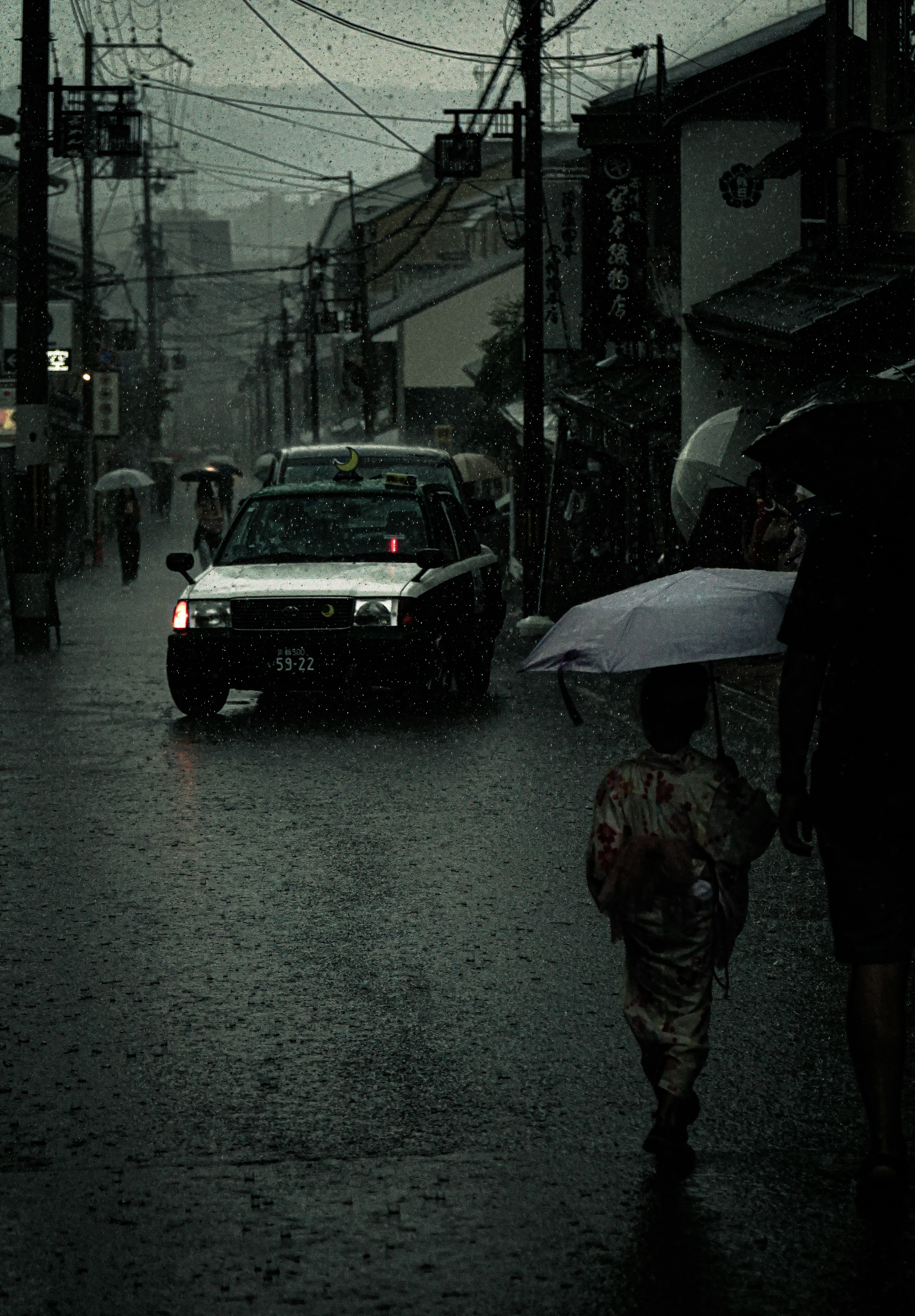 雨の中を歩く子供と傘を持つ大人の姿　暗い街並みと車が通る様子