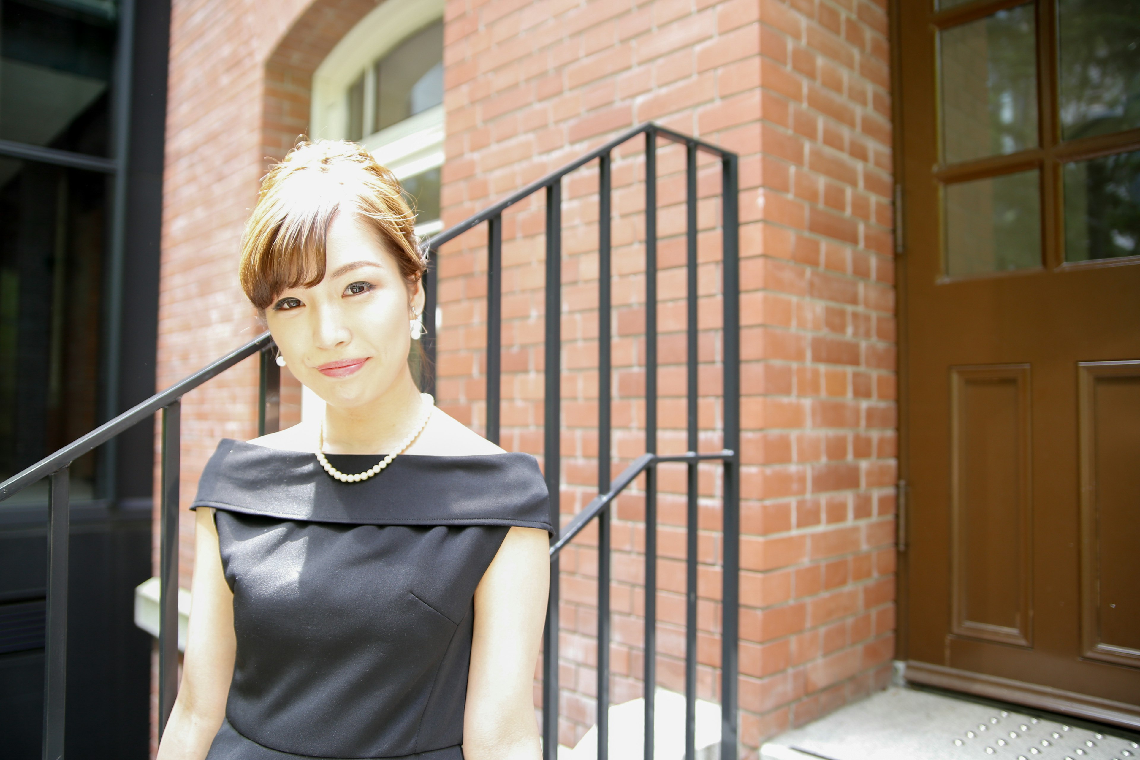 Portrait of a woman standing in front of a brick wall wearing a black dress and a pearl necklace