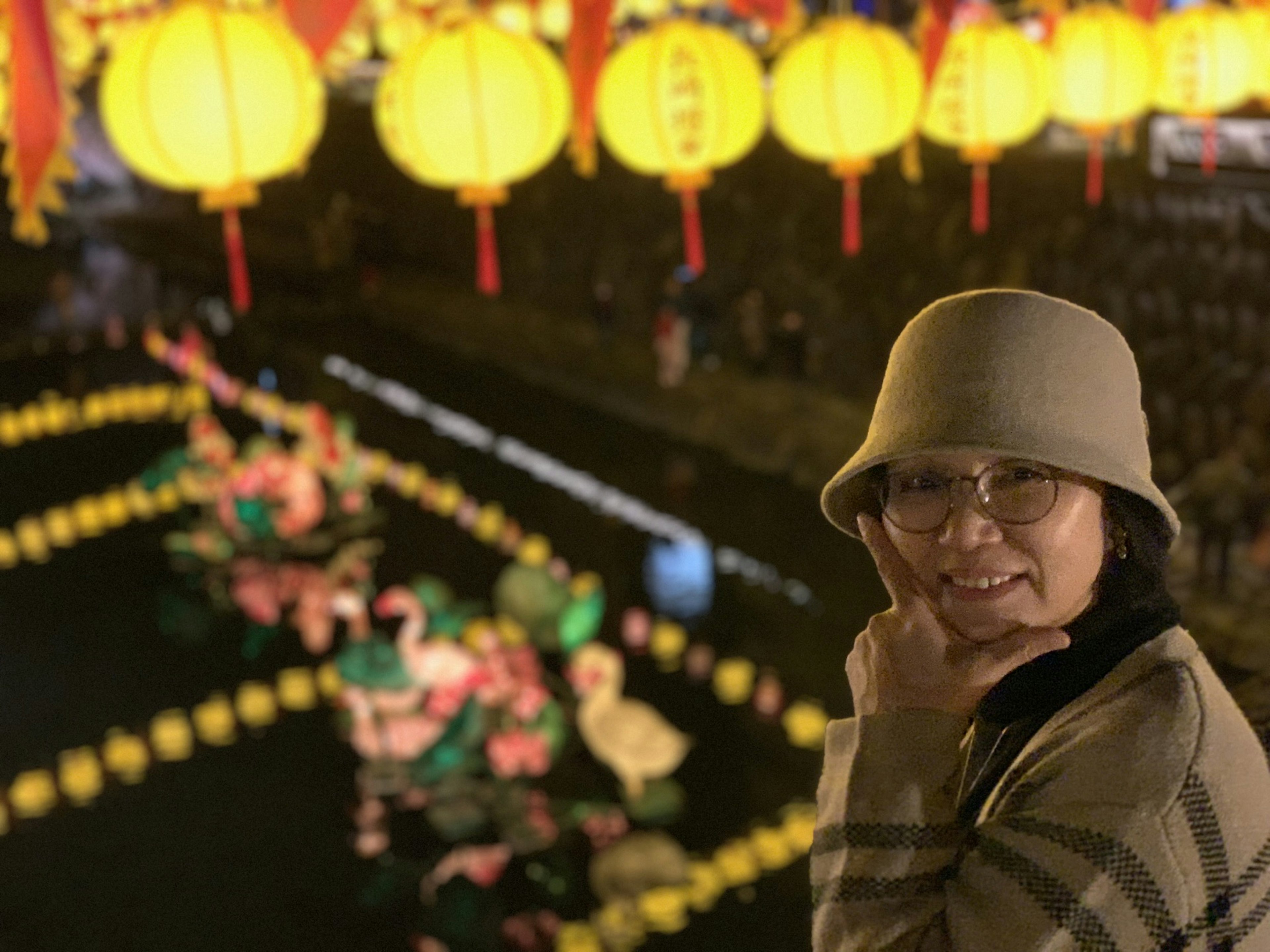 A woman smiling beside decorative ducks in water under lit lanterns at night