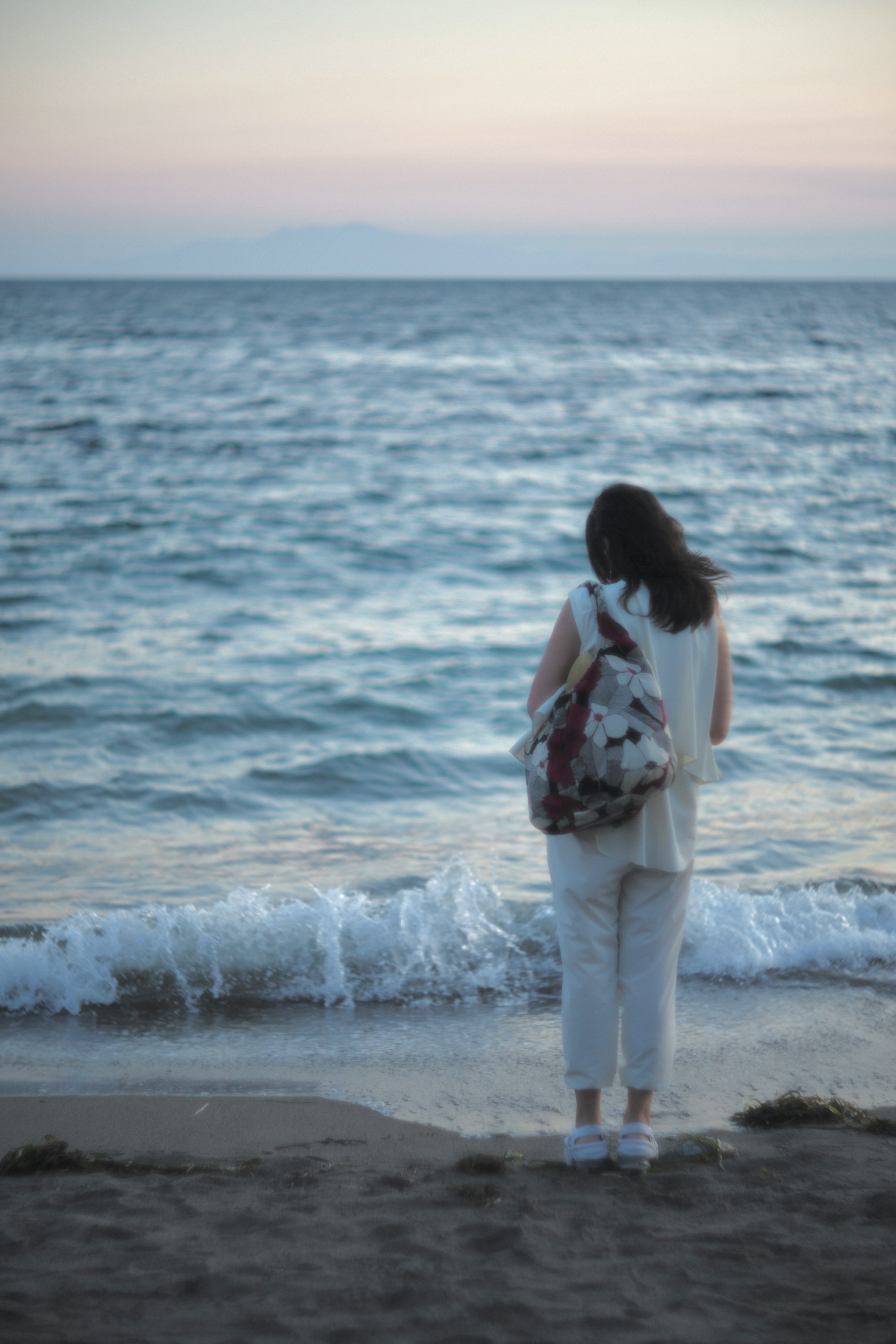 Donna in piedi sulla spiaggia che guarda le onde indossando abiti bianchi e uno zaino