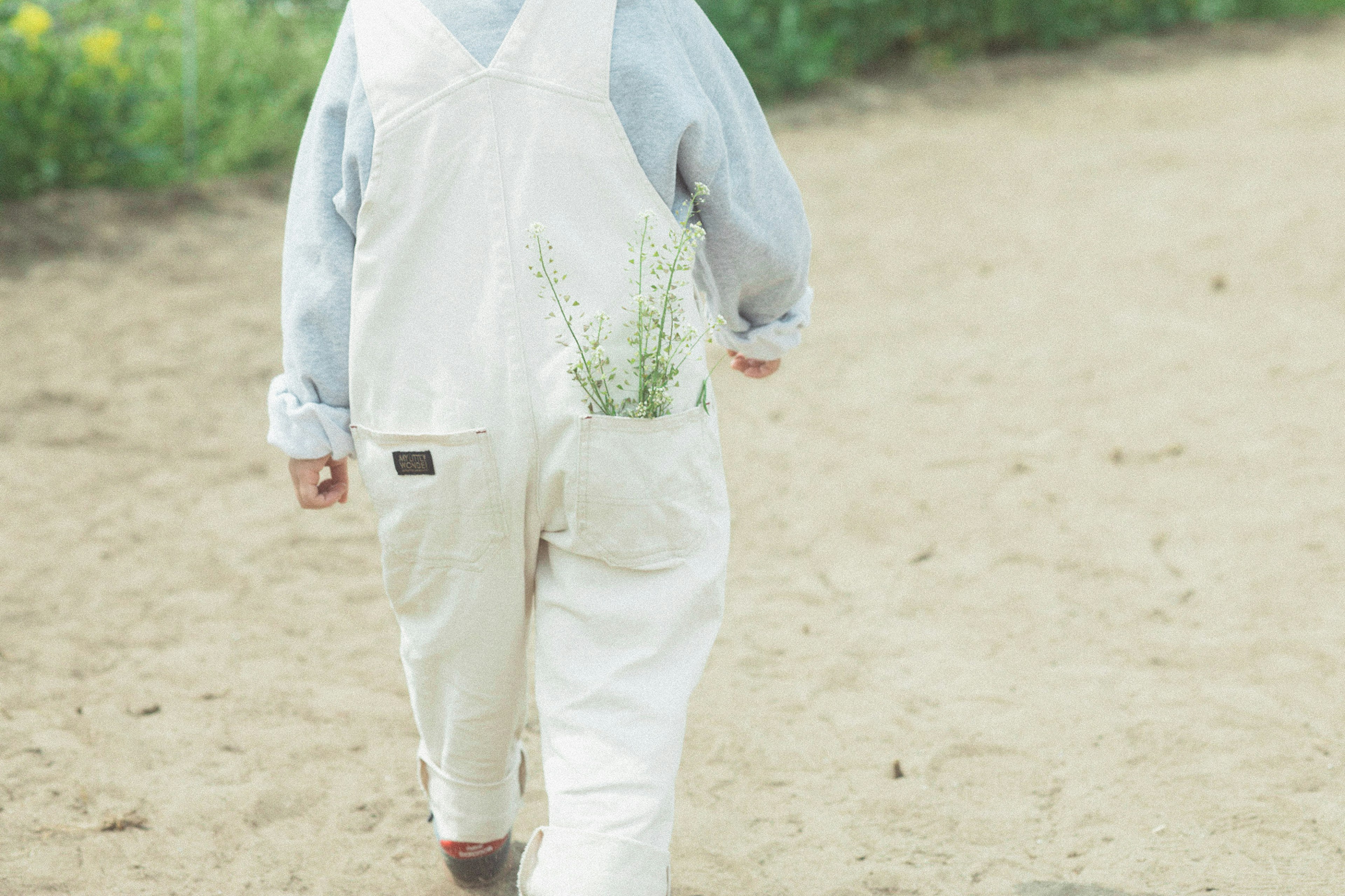 Kind in weißen Latzhosen, das auf einem sandigen Weg mit Blumen in der Tasche geht