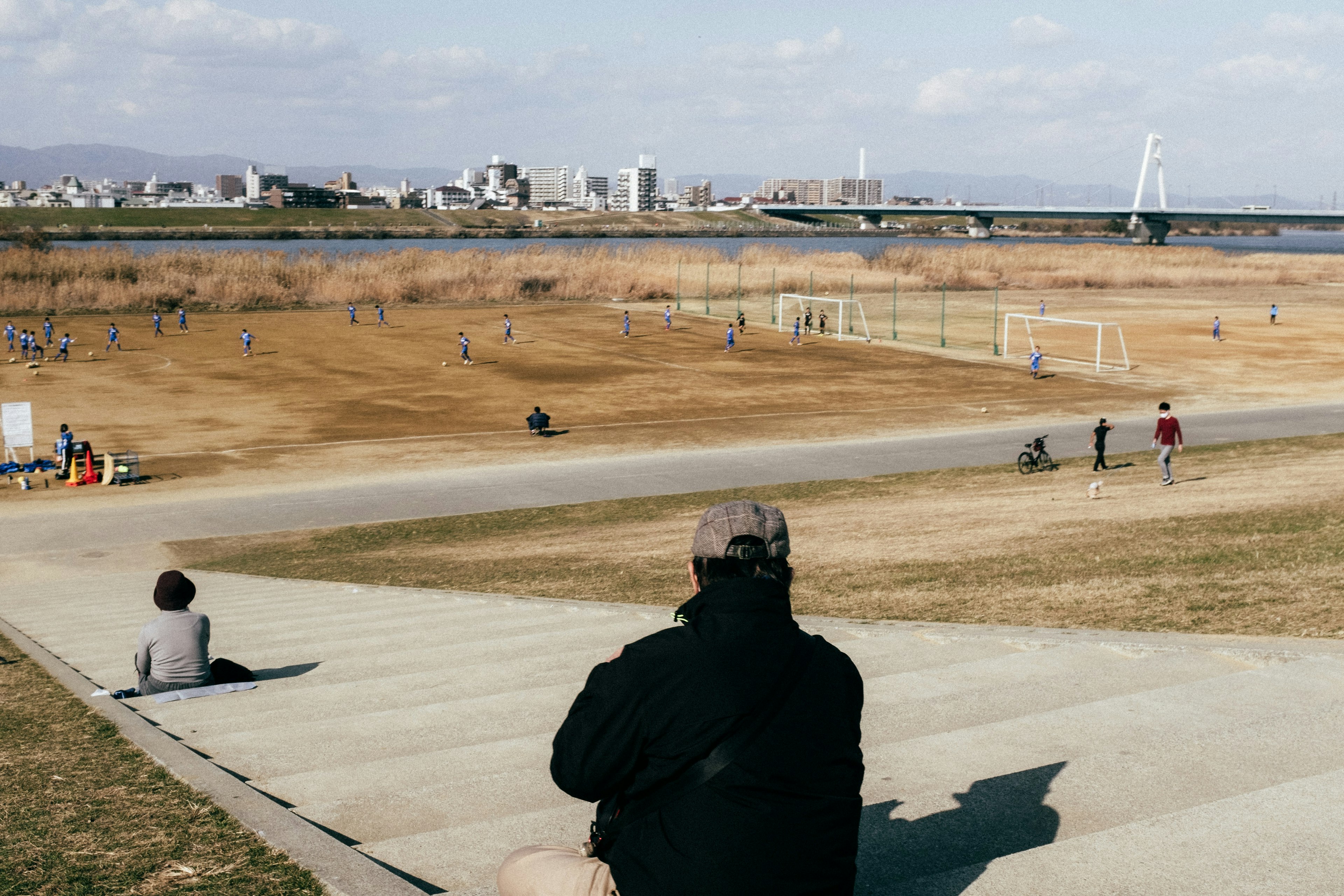 A person sitting on a sloped path overlooking a spacious riverside area with people playing