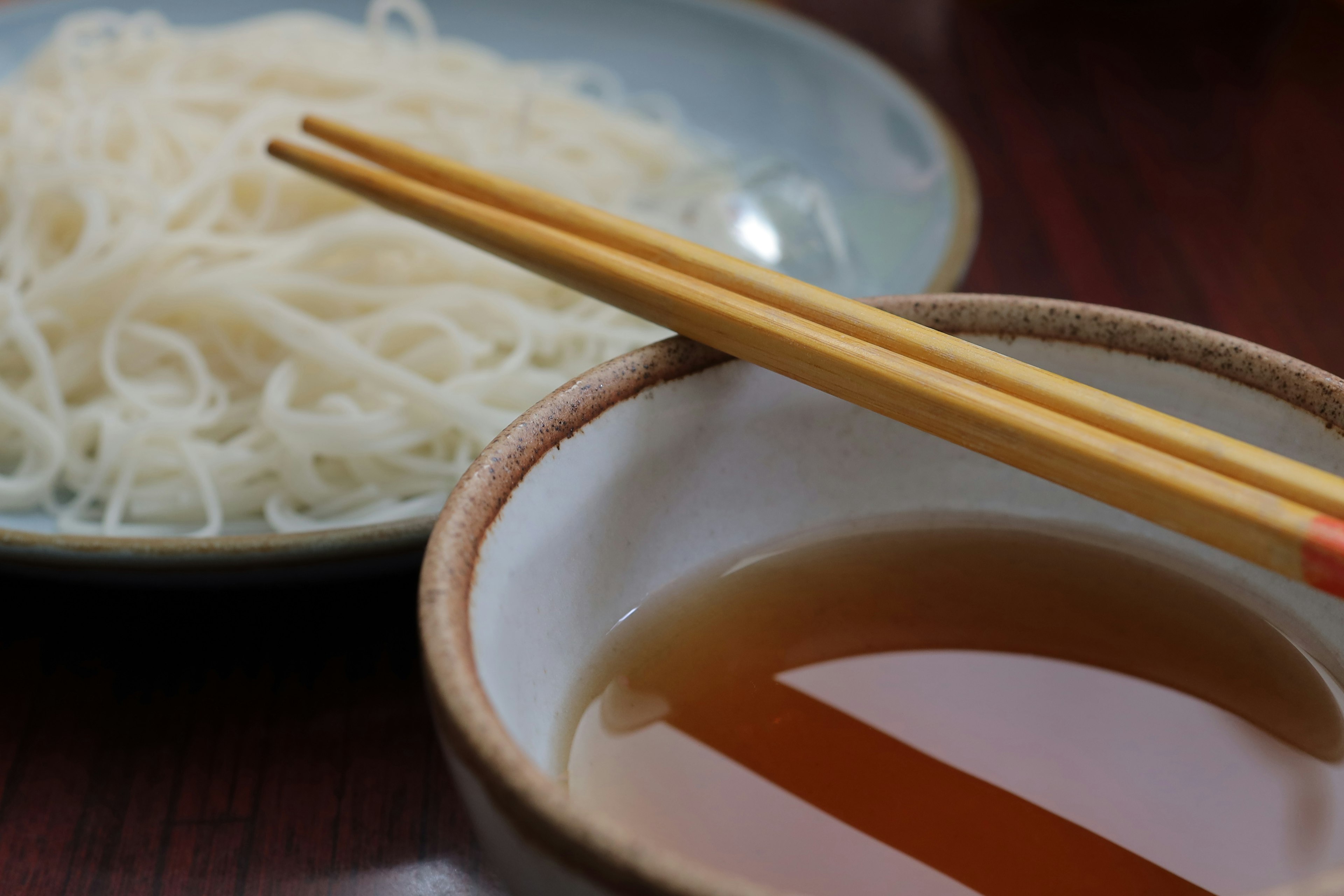 Bowl with dipping sauce and chopsticks resting next to a plate of thin noodles