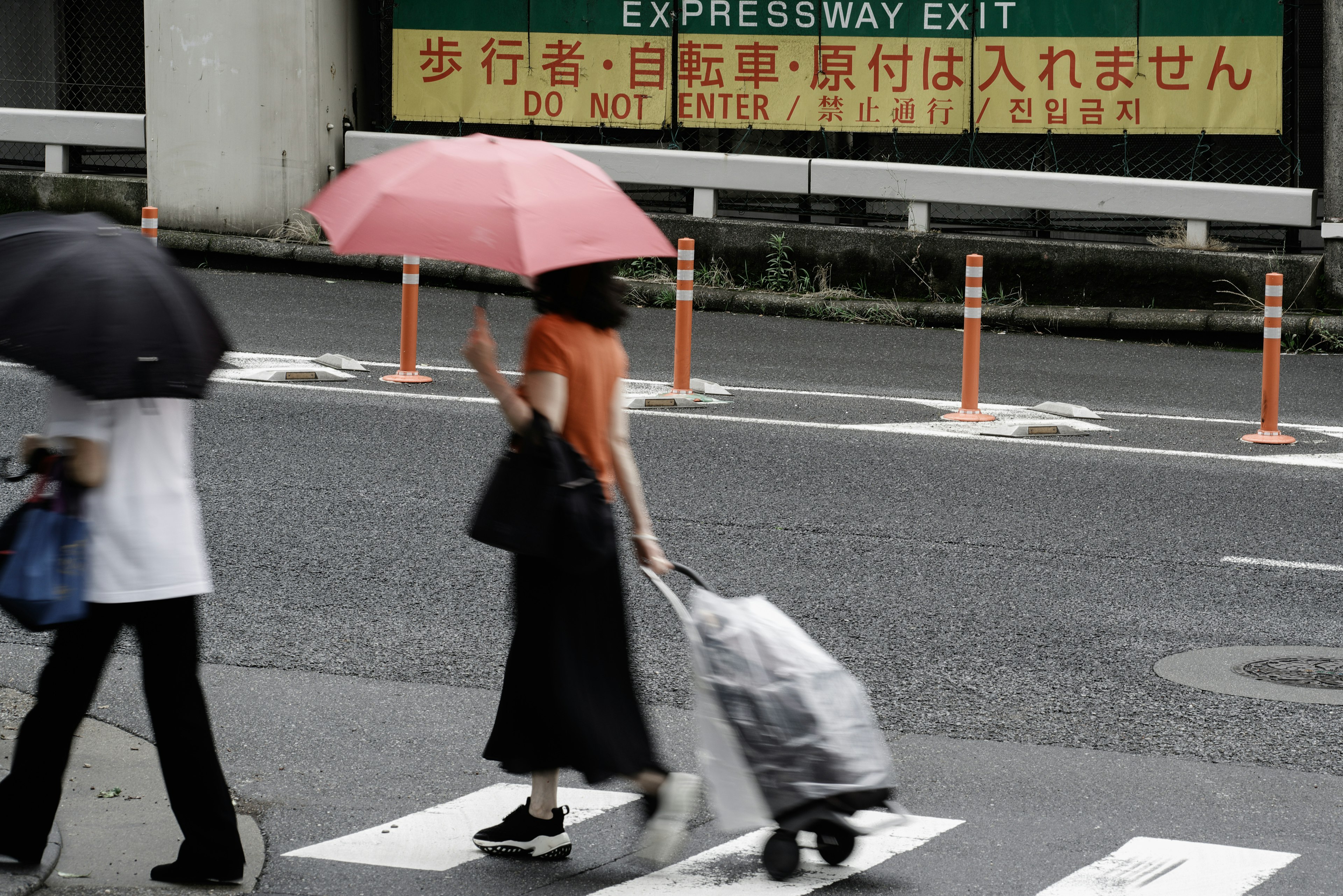 A woman with a pink umbrella crossing the street with a shopping cart