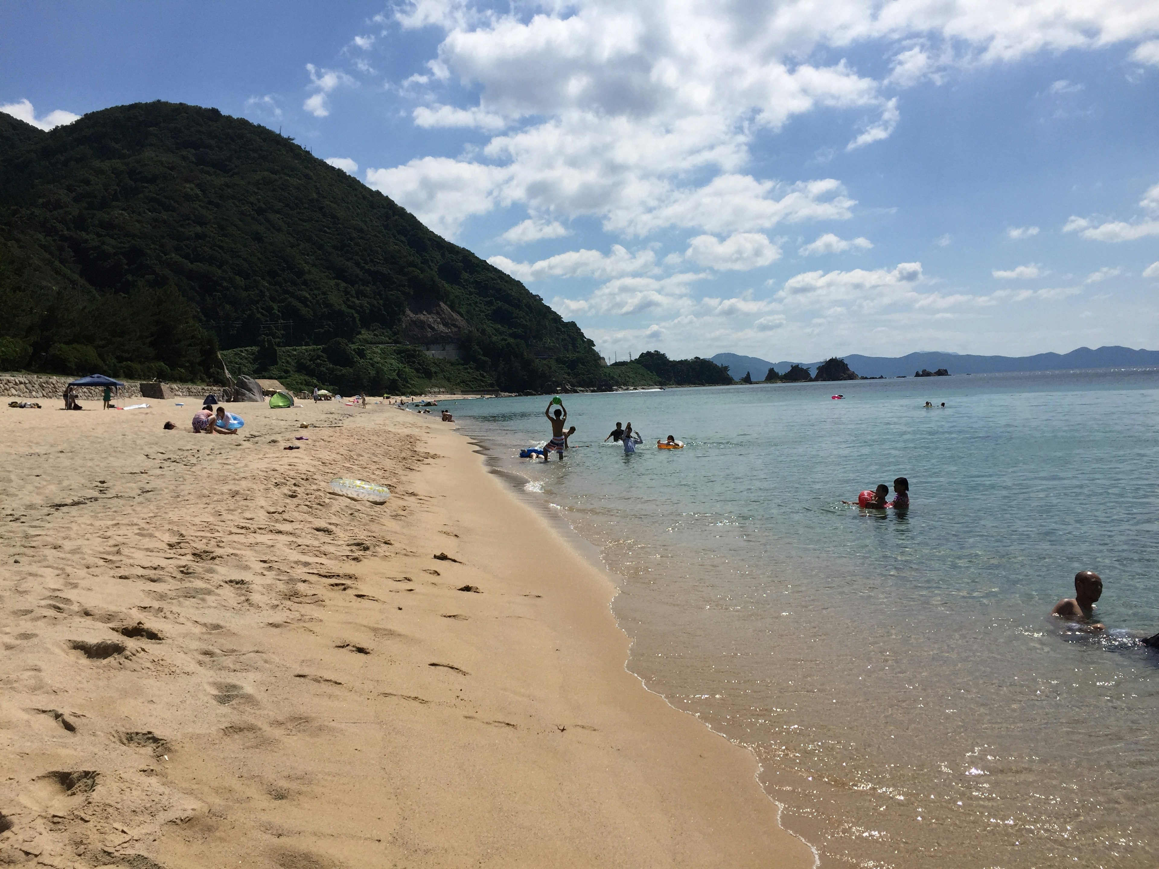 Orang-orang menikmati hari yang cerah di pantai dengan air jernih dan pasir