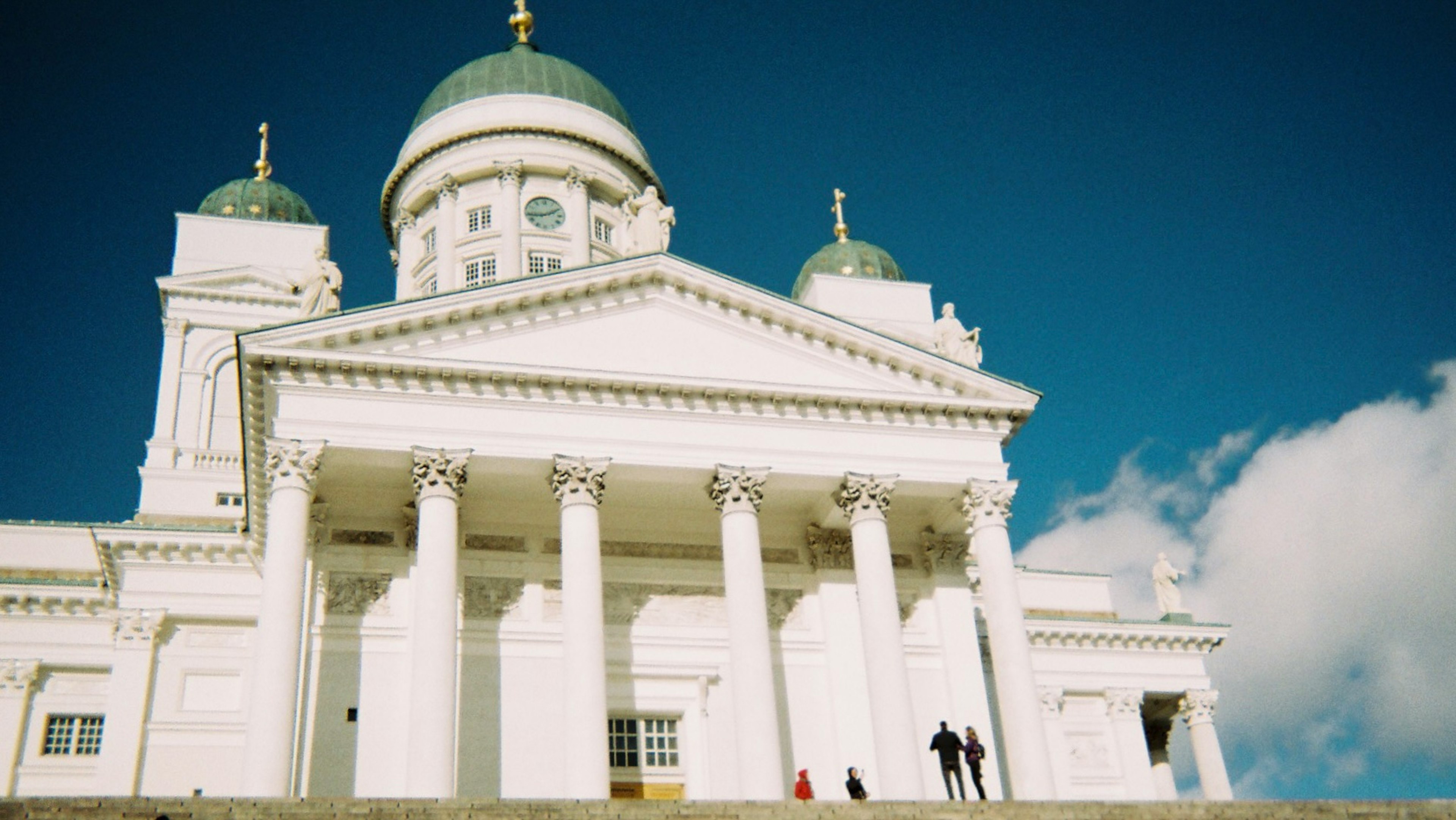 Helsinki Cathedral with white exterior and green domes under a blue sky featuring people