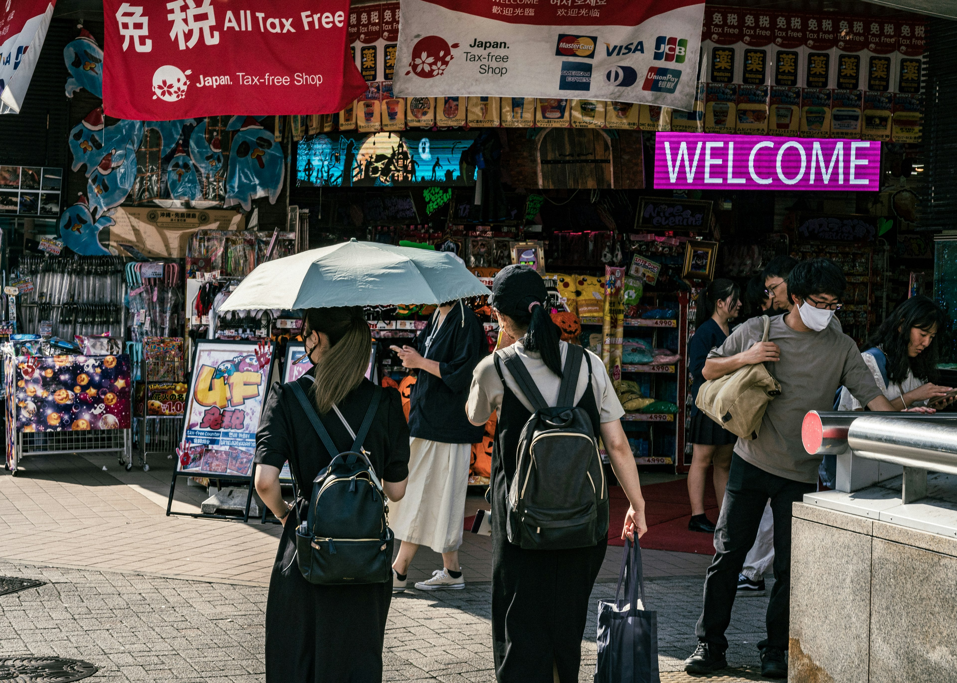 Two tourists walking with an umbrella in front of a stall featuring colorful signs