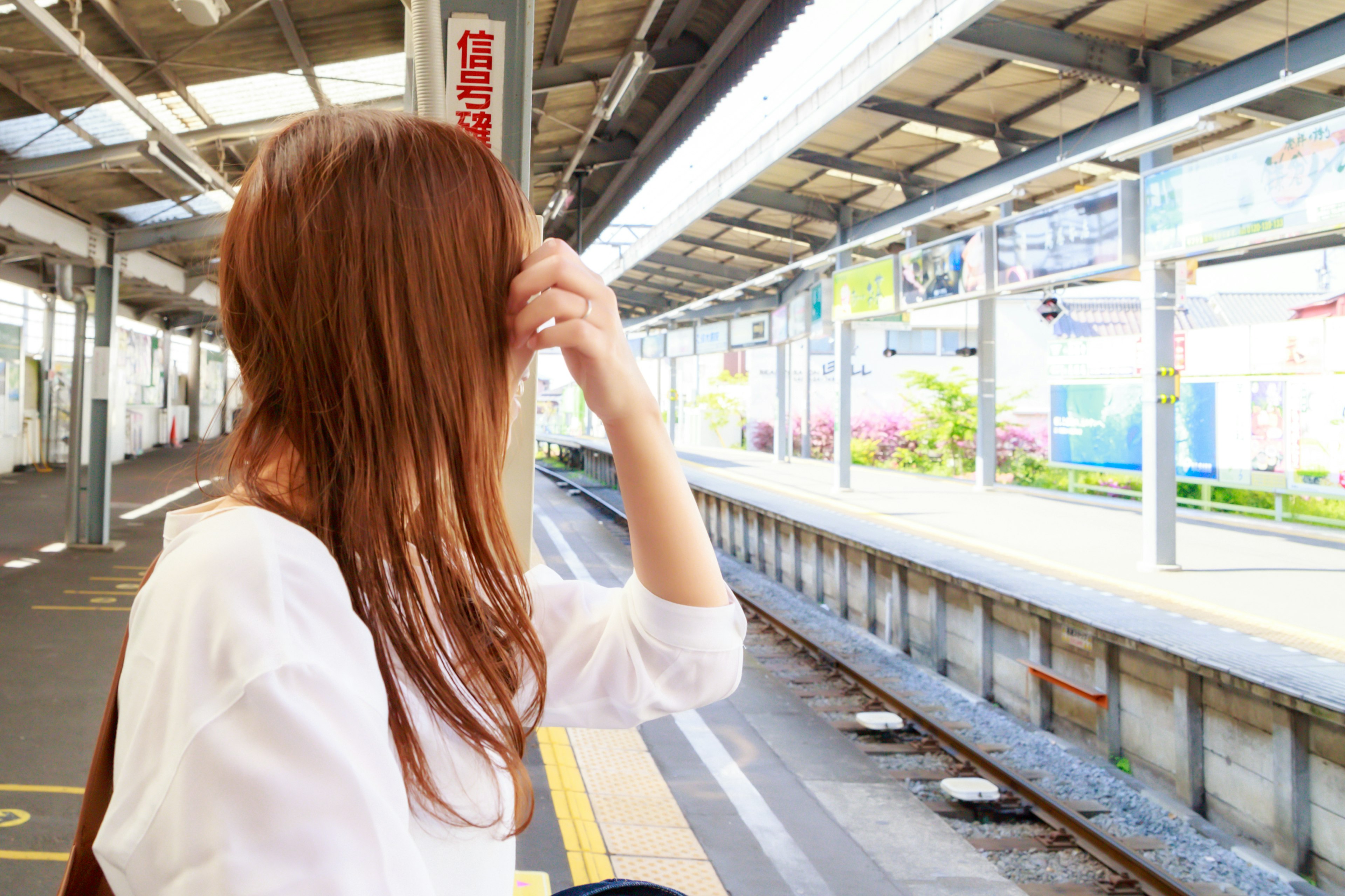 駅のホームで待つ女性の後ろ姿 髪が長くて茶色の髪を持つ 白いシャツを着ている