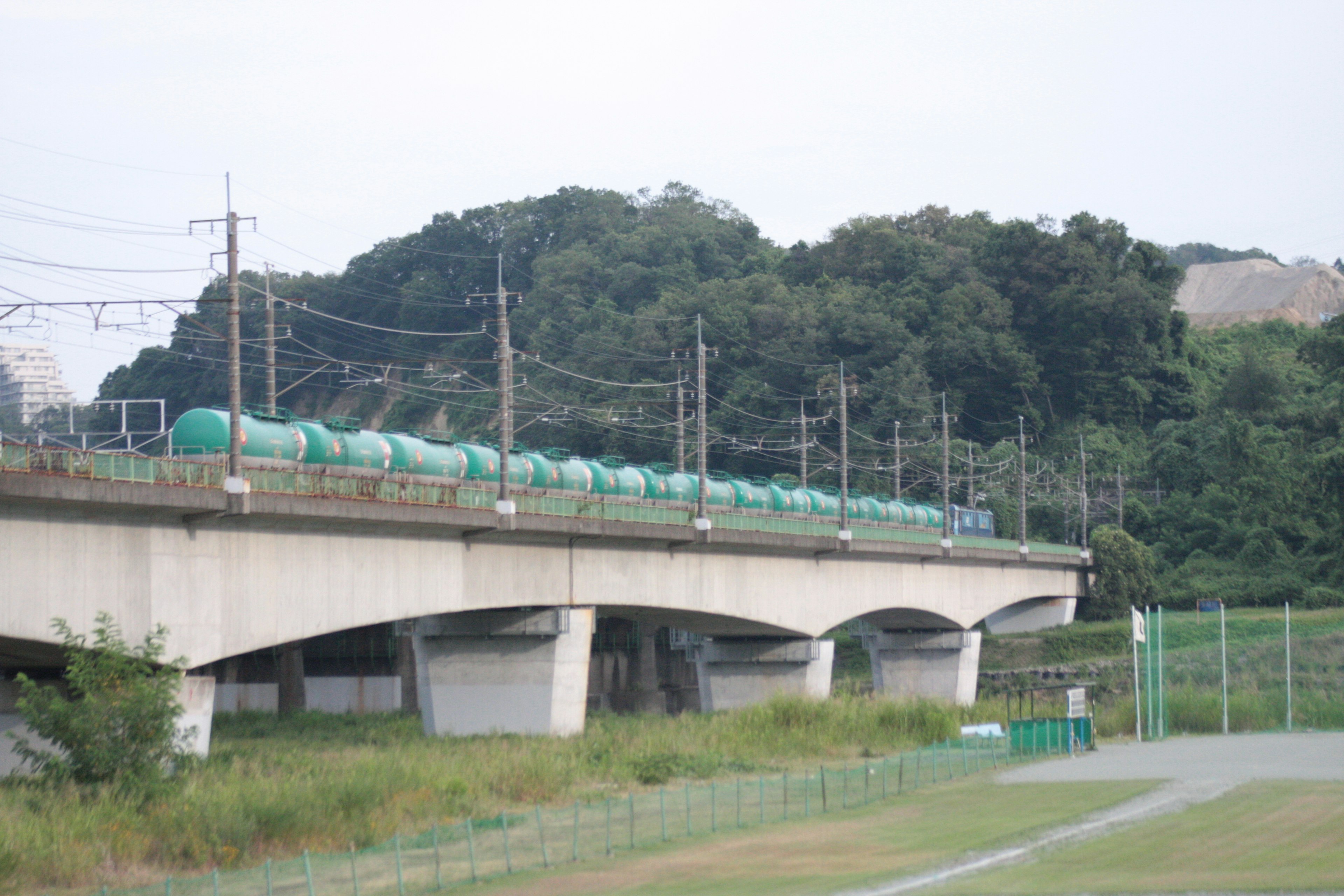 Tren que transporta tanques verdes sobre un puente con vegetación exuberante al fondo