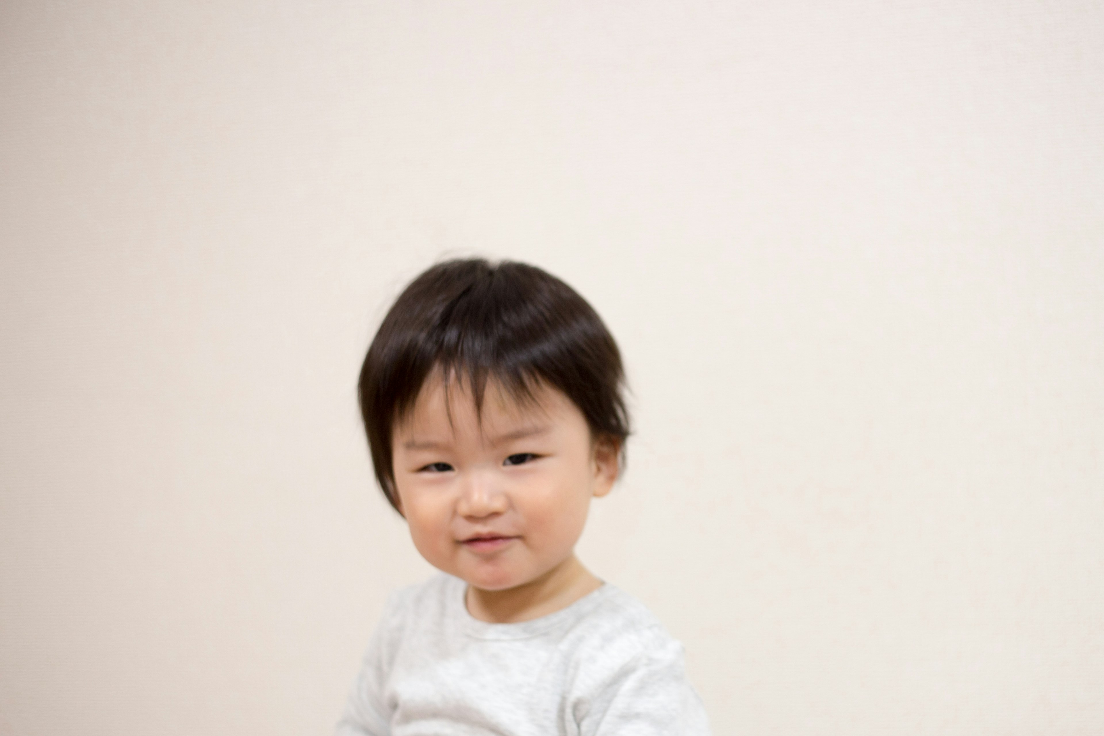 A small boy standing in front of a white background with short black hair and wearing a gray outfit