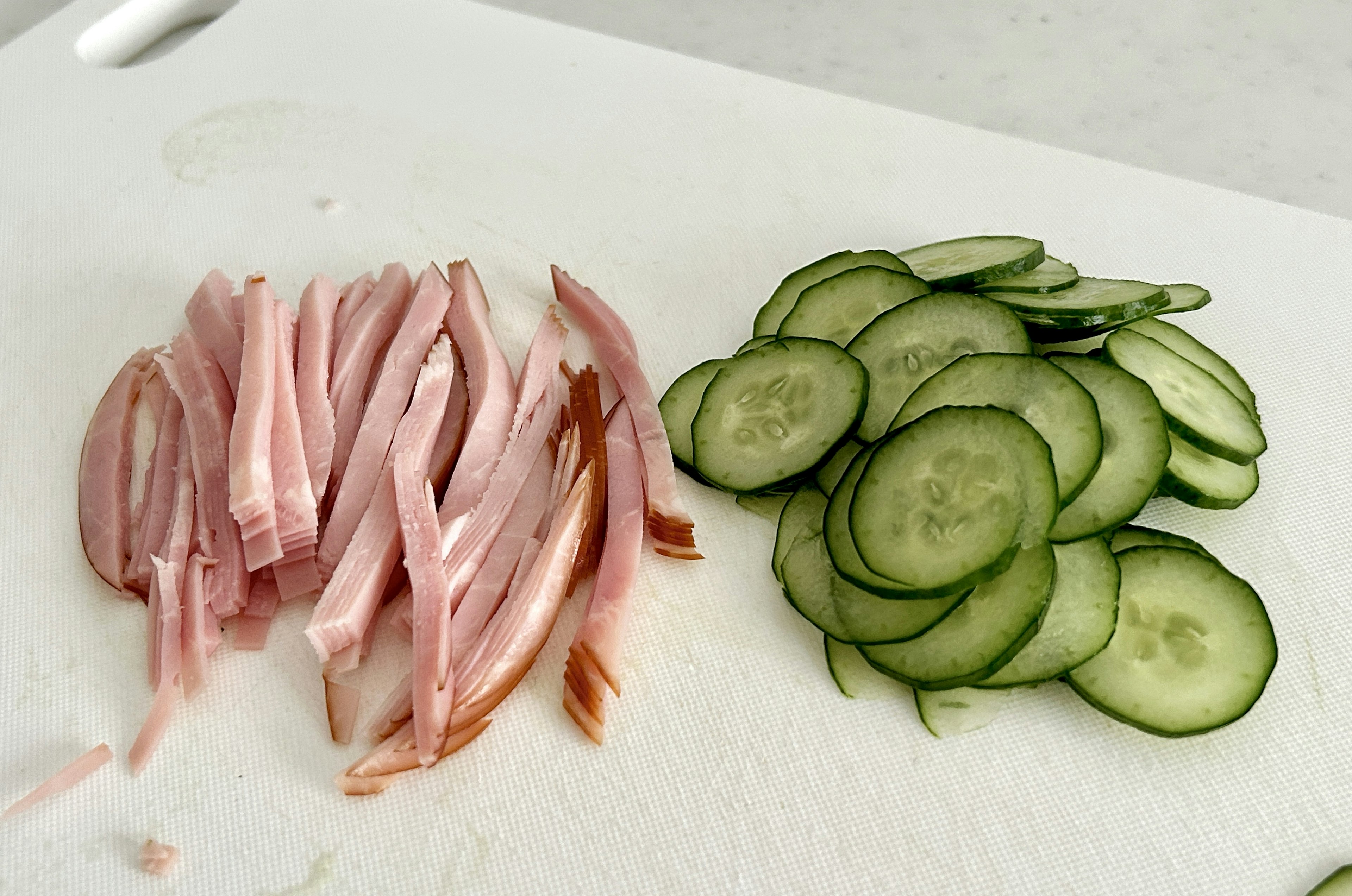 Sliced cucumbers and pink ham arranged on a white cutting board