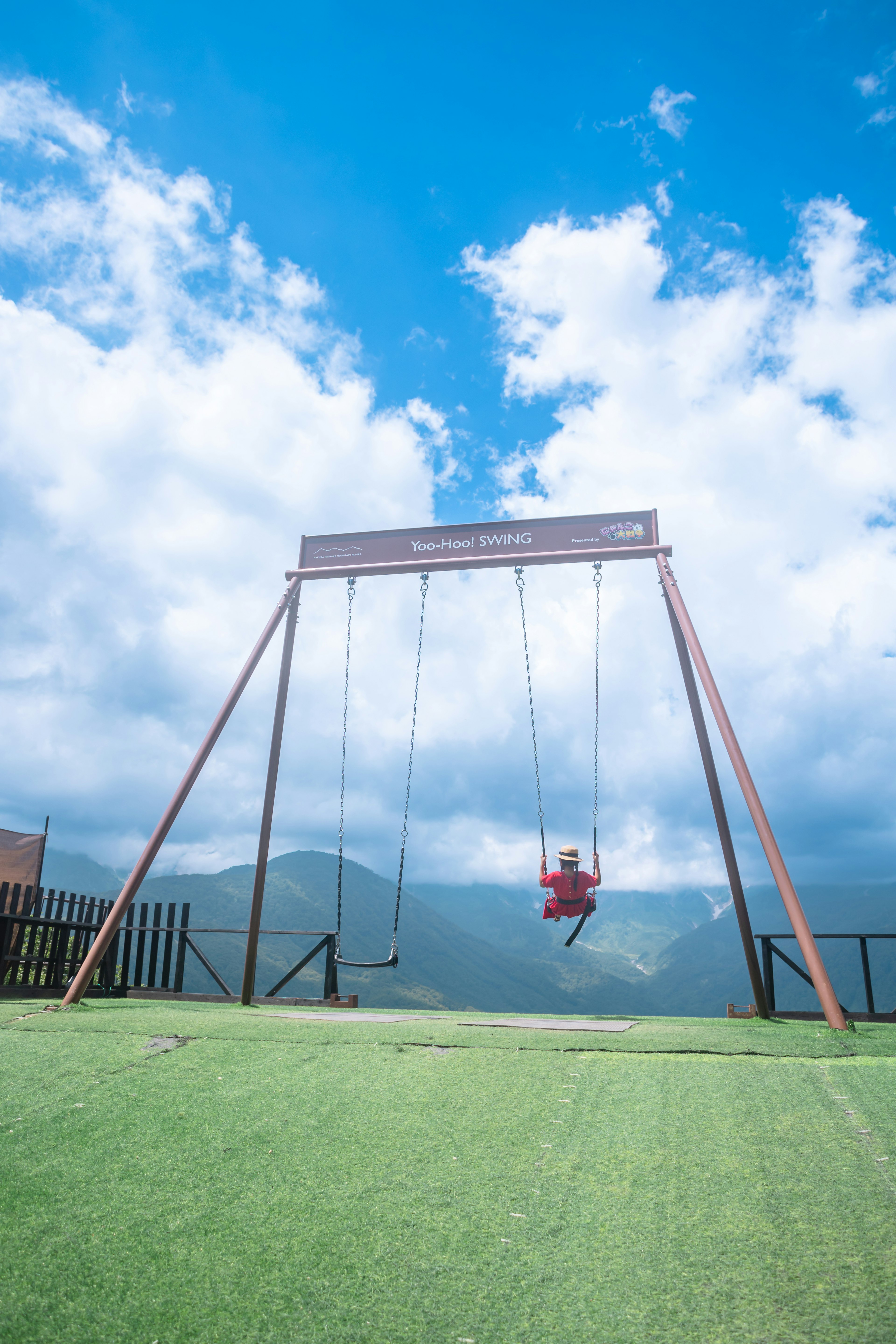 青い空と雲の下にある赤いブランコと緑の芝生