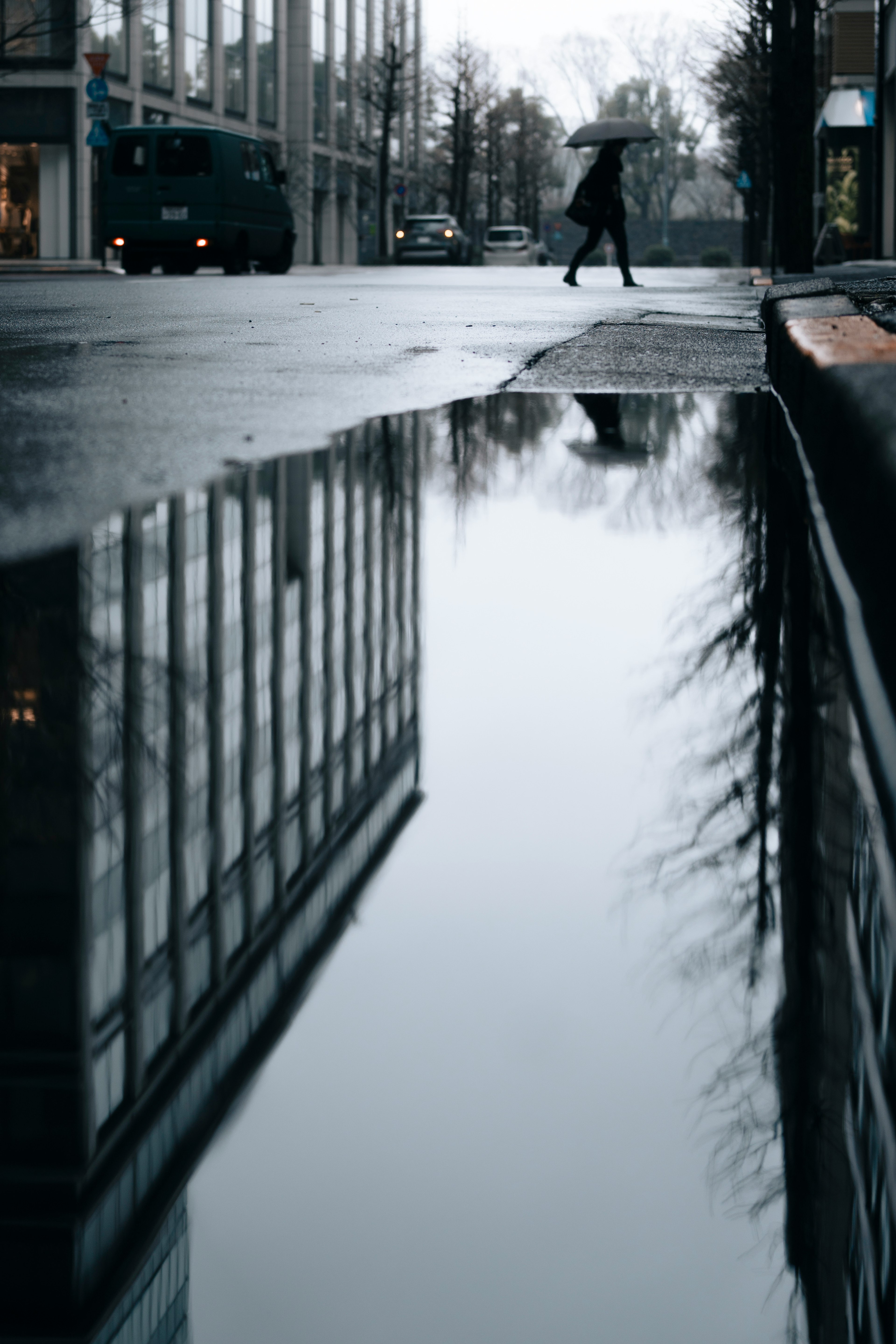 雨上がりの街角に映るビルと木々の反射を持つ水たまりを歩く人