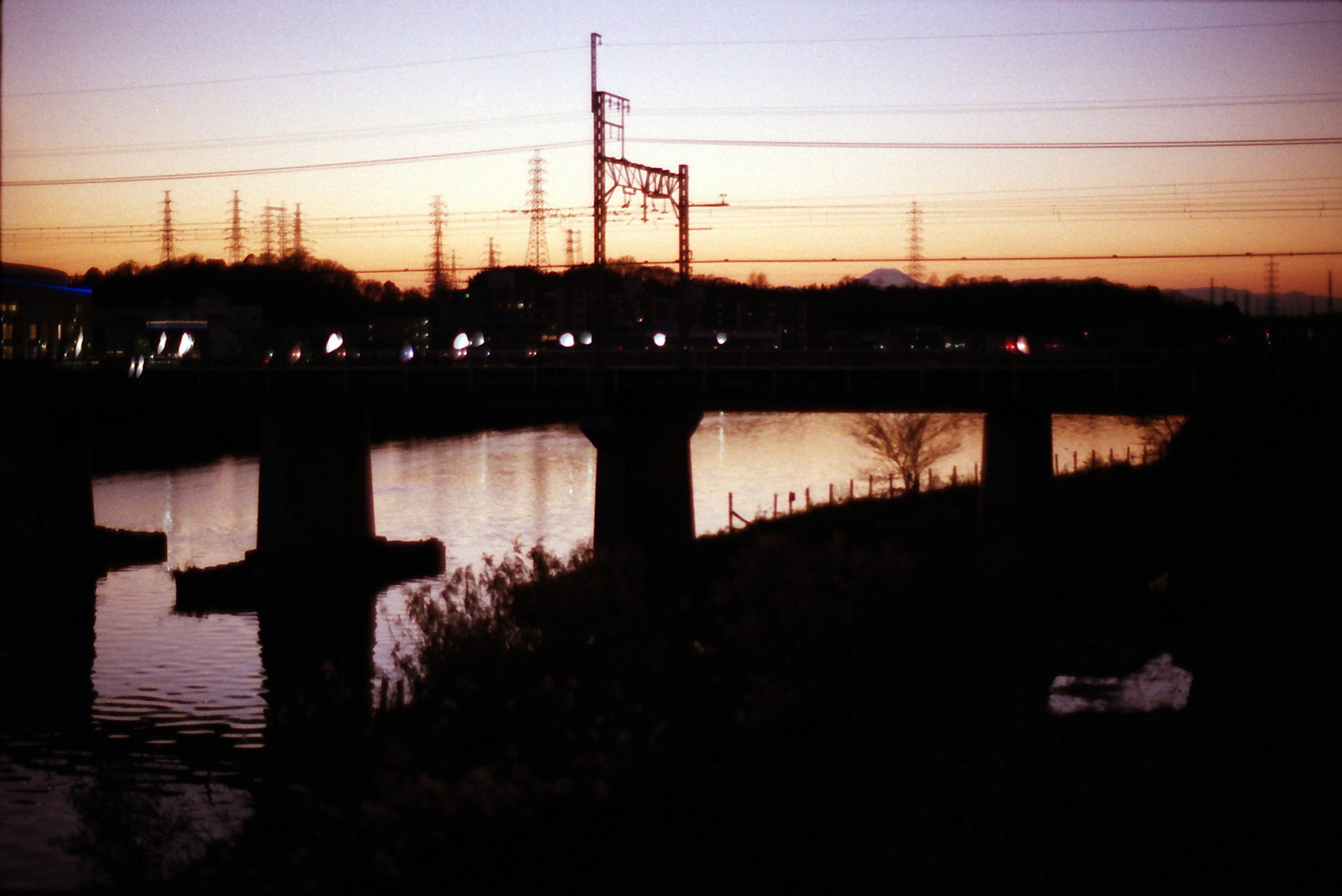 Dämmerungsansicht eines Flusses und einer Brücke mit sichtbaren Stromleitungen und Eisenbahn